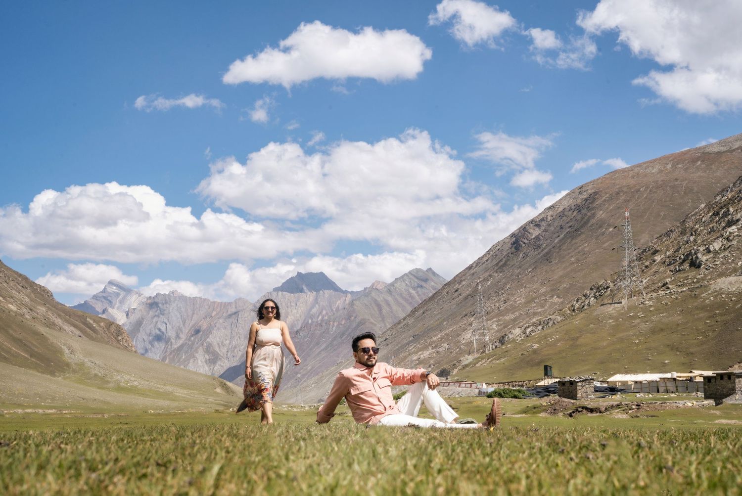 Couple relaxing on green meadow with stunning mountain backdrop under blue sky in a scenic valley.
