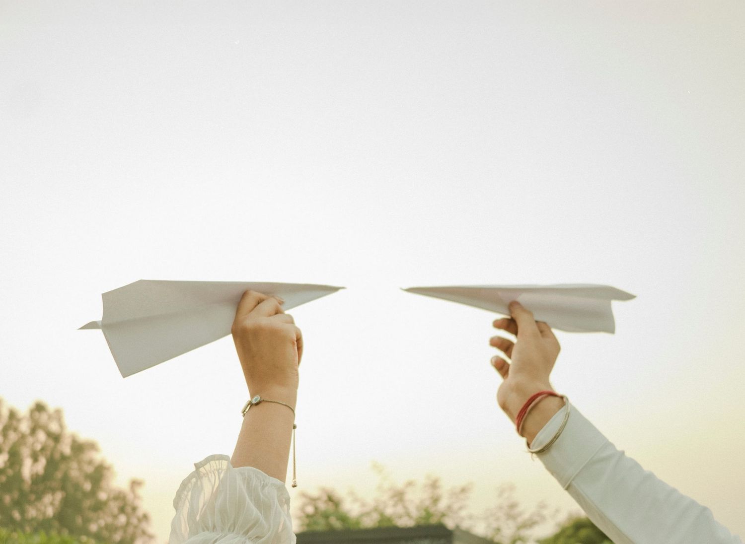Two hands holding white paper airplanes up against a bright sky outdoors.