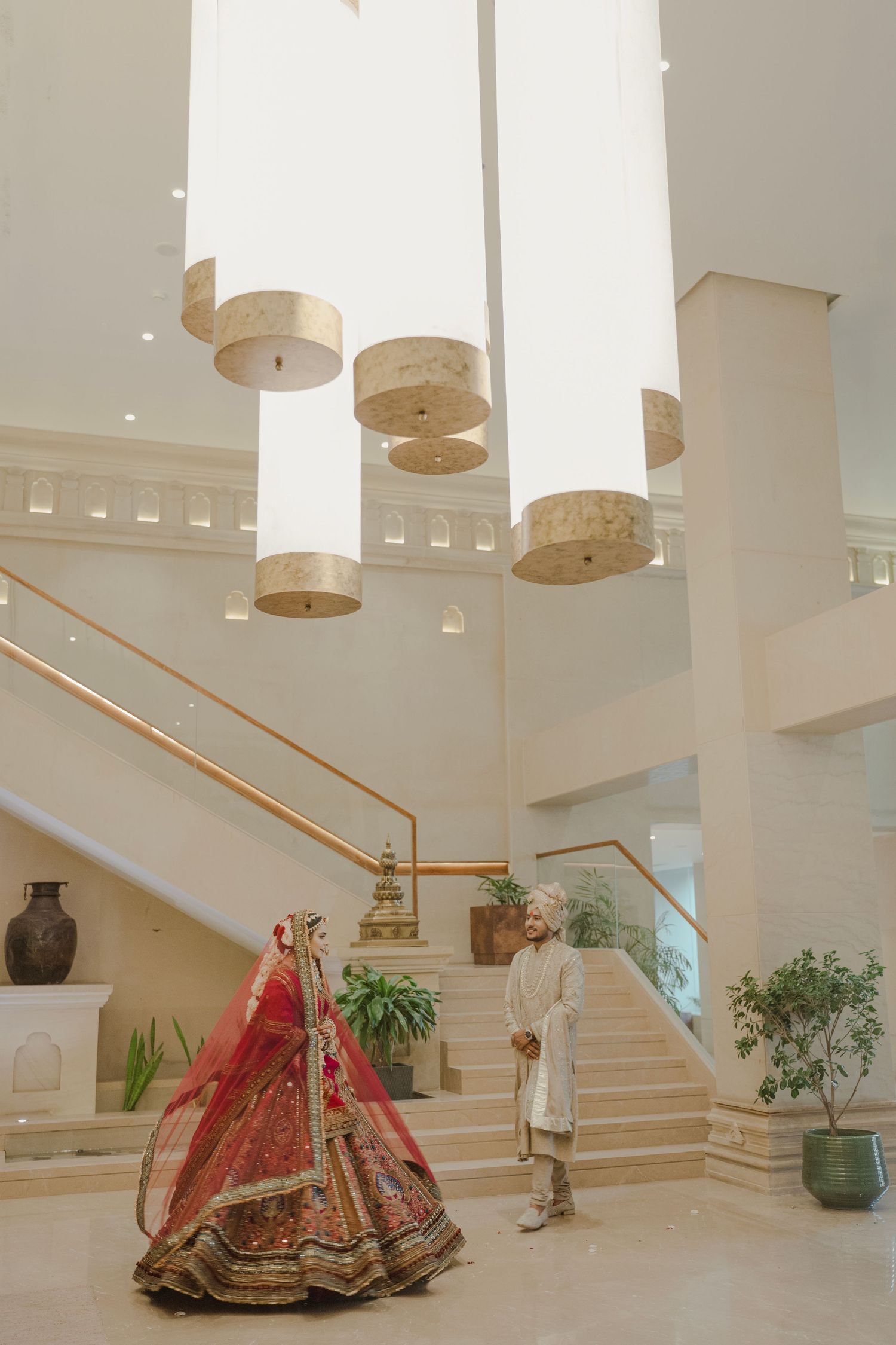 Indian bride in red lehenga and groom in sherwani stand in elegant white wedding venue with grand chandeliers and staircase.