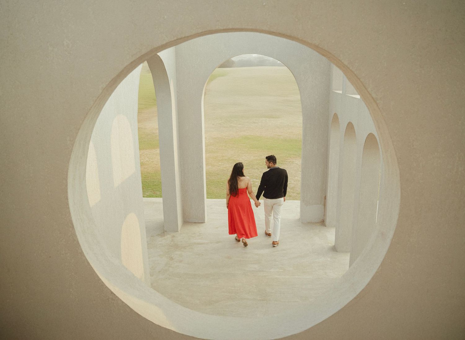 Couple in red dress and black outfit framed by circular white arch with colonnades and scenic landscape.