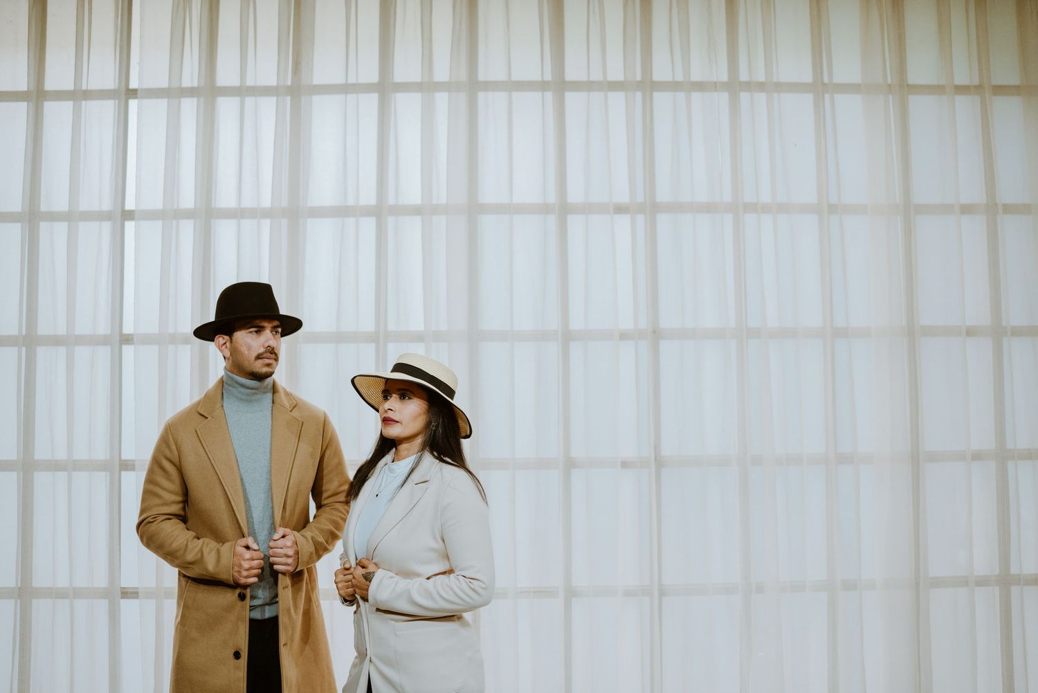 Stylish couple posing outdoors, man in brown coat and black hat, woman in white blazer and straw hat.