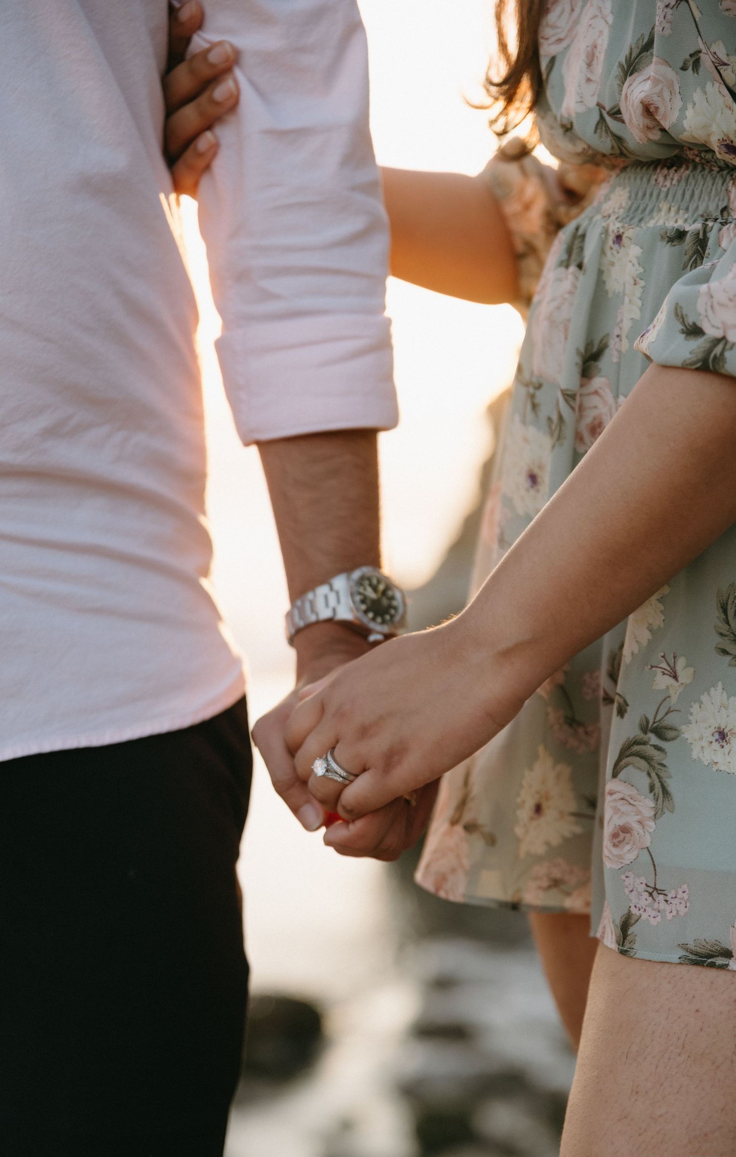 Couple holding hands outdoors at sunset, woman wearing floral dress and ring, man wearing white shirt and silver watch.