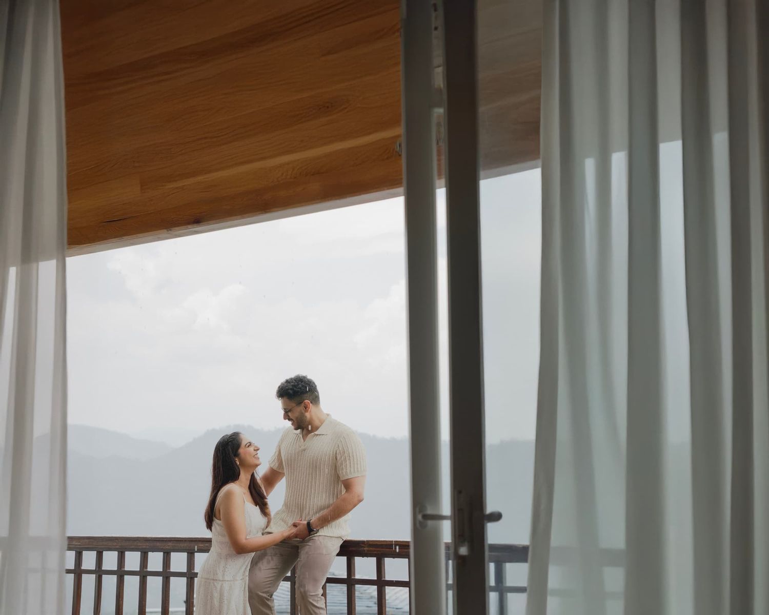 Couple sharing a romantic moment on a mountain view balcony, dressed in elegant neutral tones.