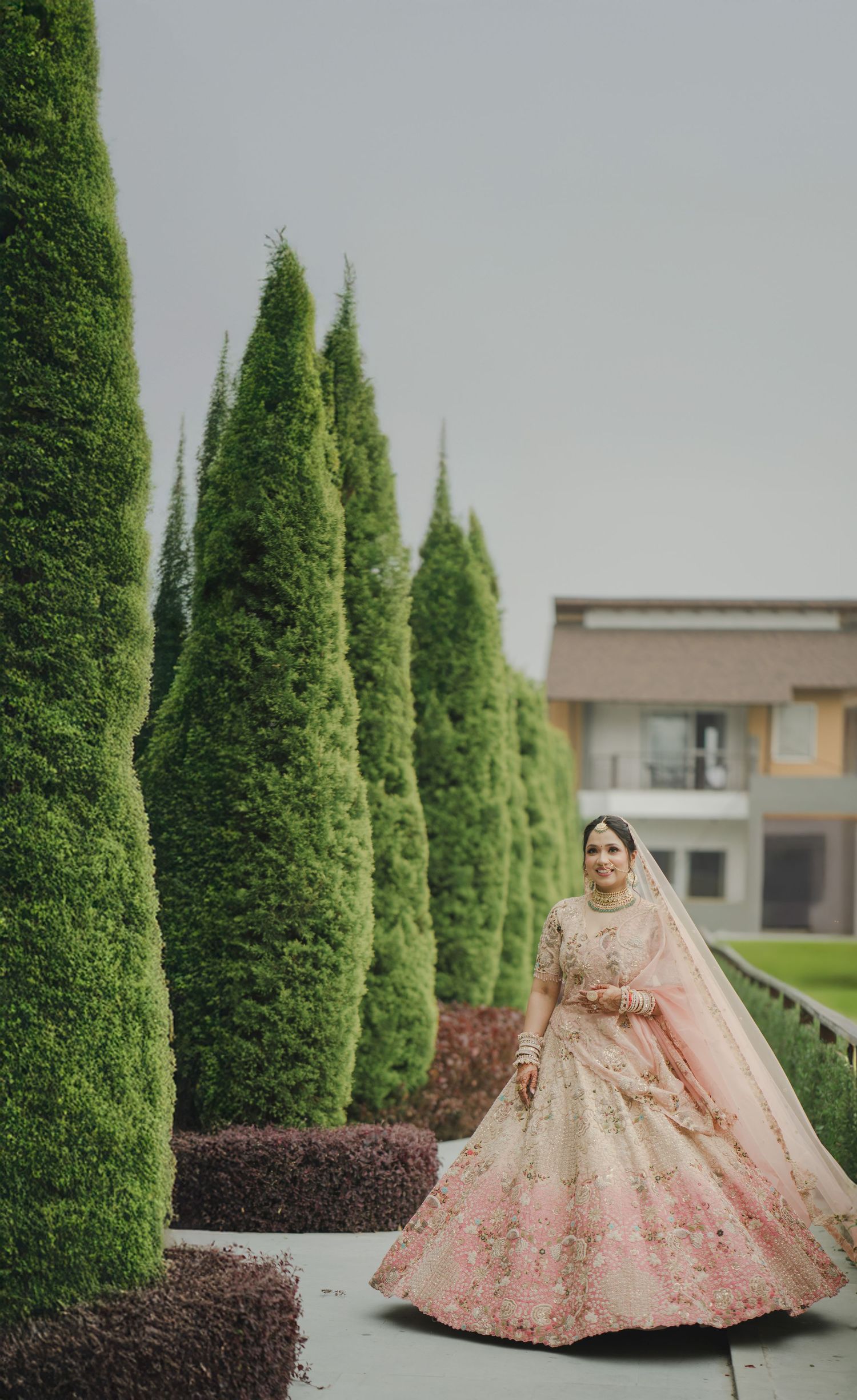 A bride in a pink embroidered lehenga poses elegantly beside tall manicured cypress trees in a garden setting.