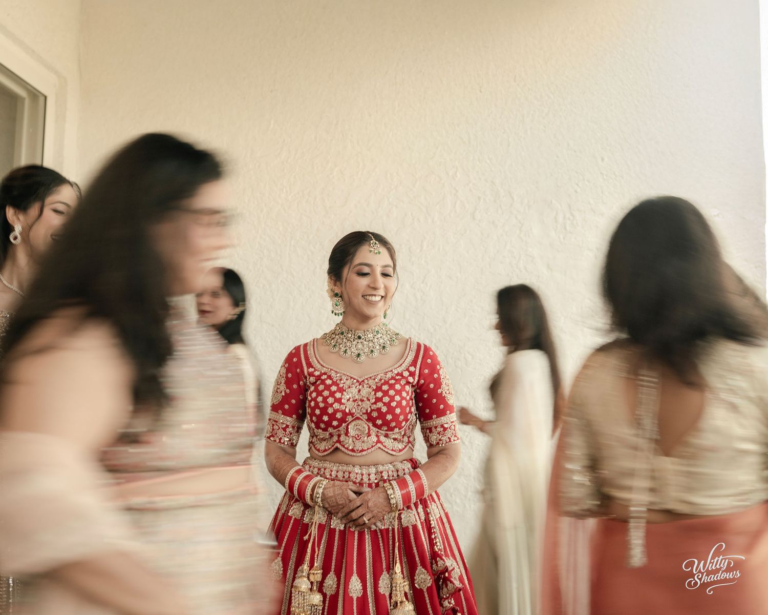 Bride in red lehenga with gold jewelry smiles as bridesmaids in white blur around her at Indian wedding.