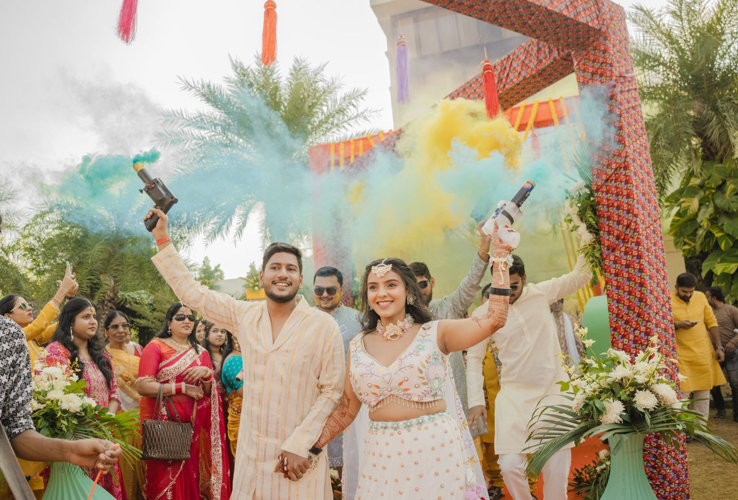 Indian couple holding colorful smoke bombs at wedding entrance, guests celebrating in background with vibrant traditional attire.