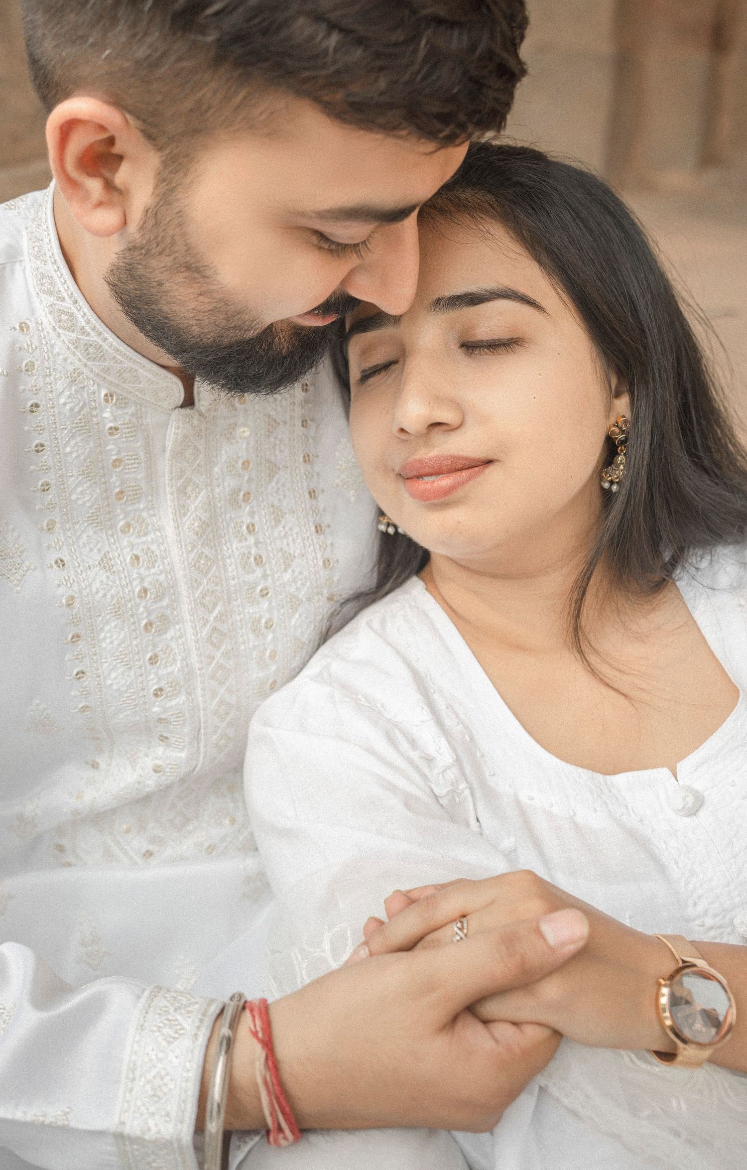 A couple in white traditional Indian attire share a tender moment, holding hands with eyes closed.