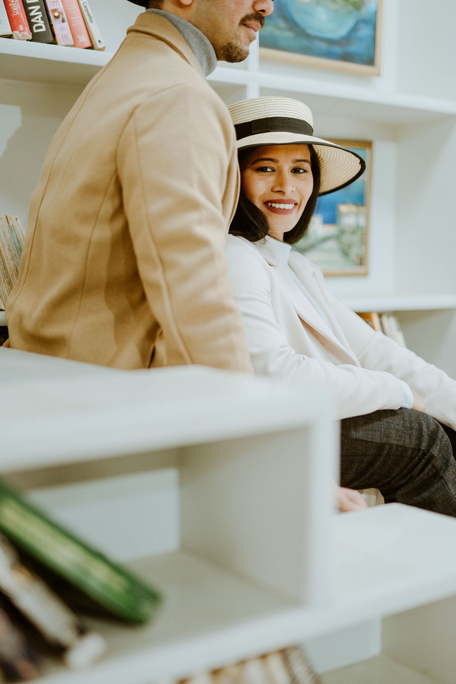 A smiling woman in a straw hat and white blouse sits beside a man in a beige blazer near a white bookshelf.
