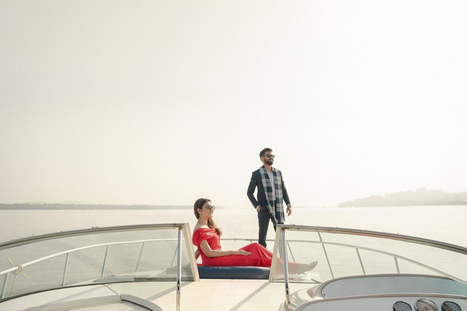 A couple relaxing on the bow of a luxury yacht on calm waters, woman in red dress seated, man standing in plaid shirt.