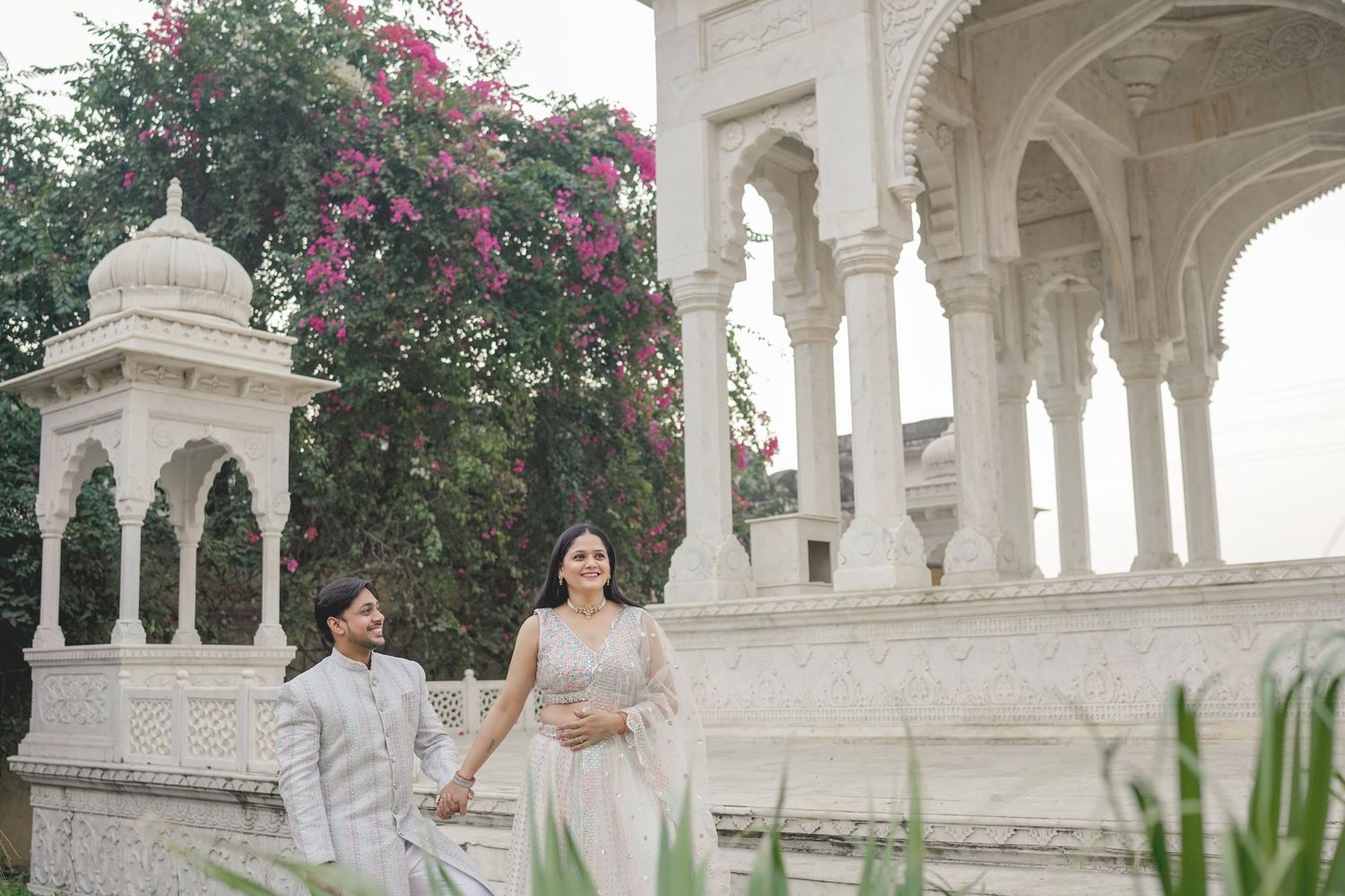 Couple in traditional Indian attire walking hand in hand at a white marble pavilion with pink bougainvillea.