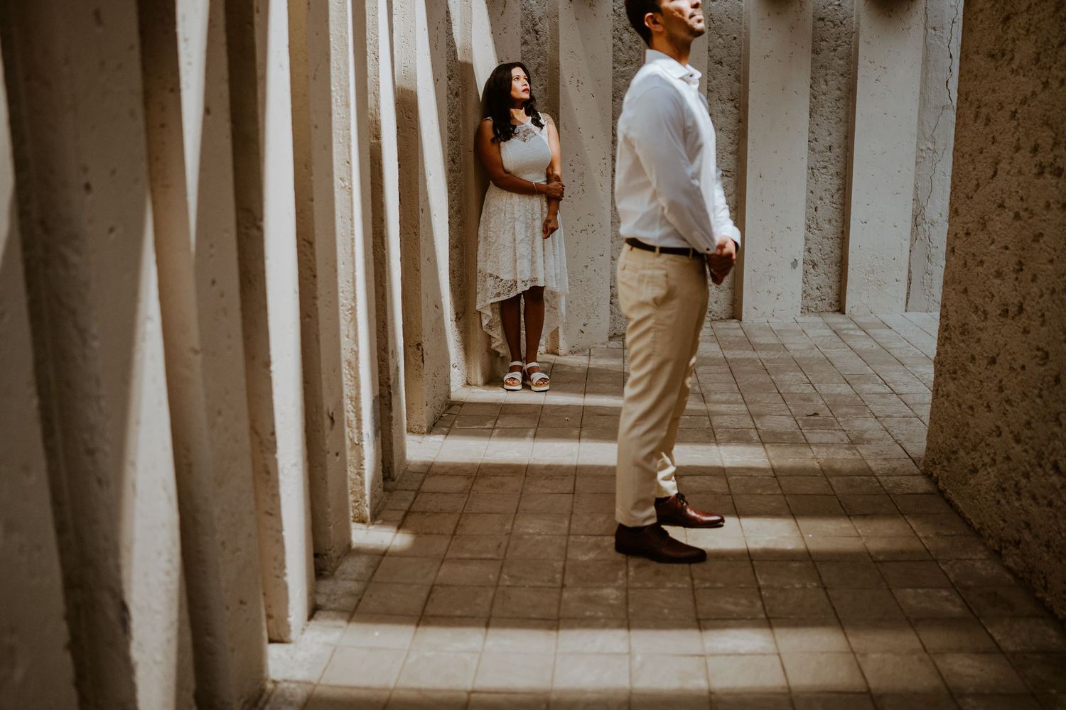 Couple standing apart between stone columns in a tiled corridor, woman in white dress, man in white shirt and khakis.