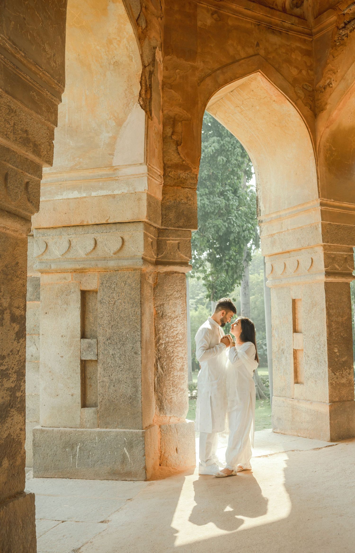 Couple in white attire embracing under ornate stone arches of an ancient Indian monument in golden sunlight.
