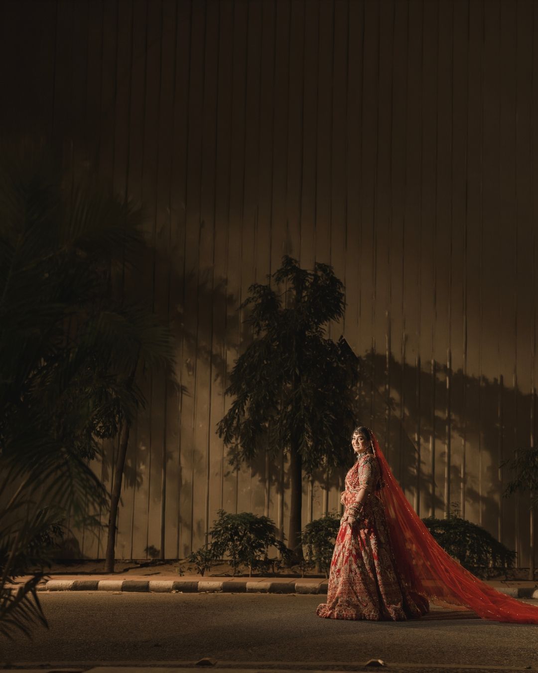 Bride in red bridal lehenga with flowing dupatta stands outdoors at night, illuminated against dark backdrop with palm trees.