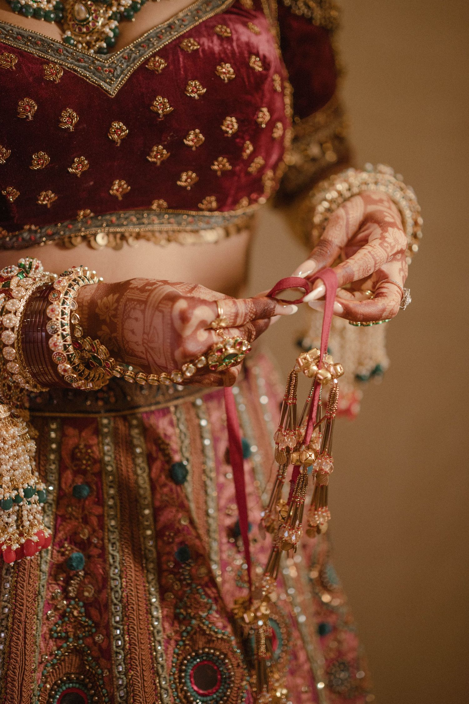 Indian bride in red lehenga with gold embroidery holds jewelry, hands adorned with mehndi and bangles.