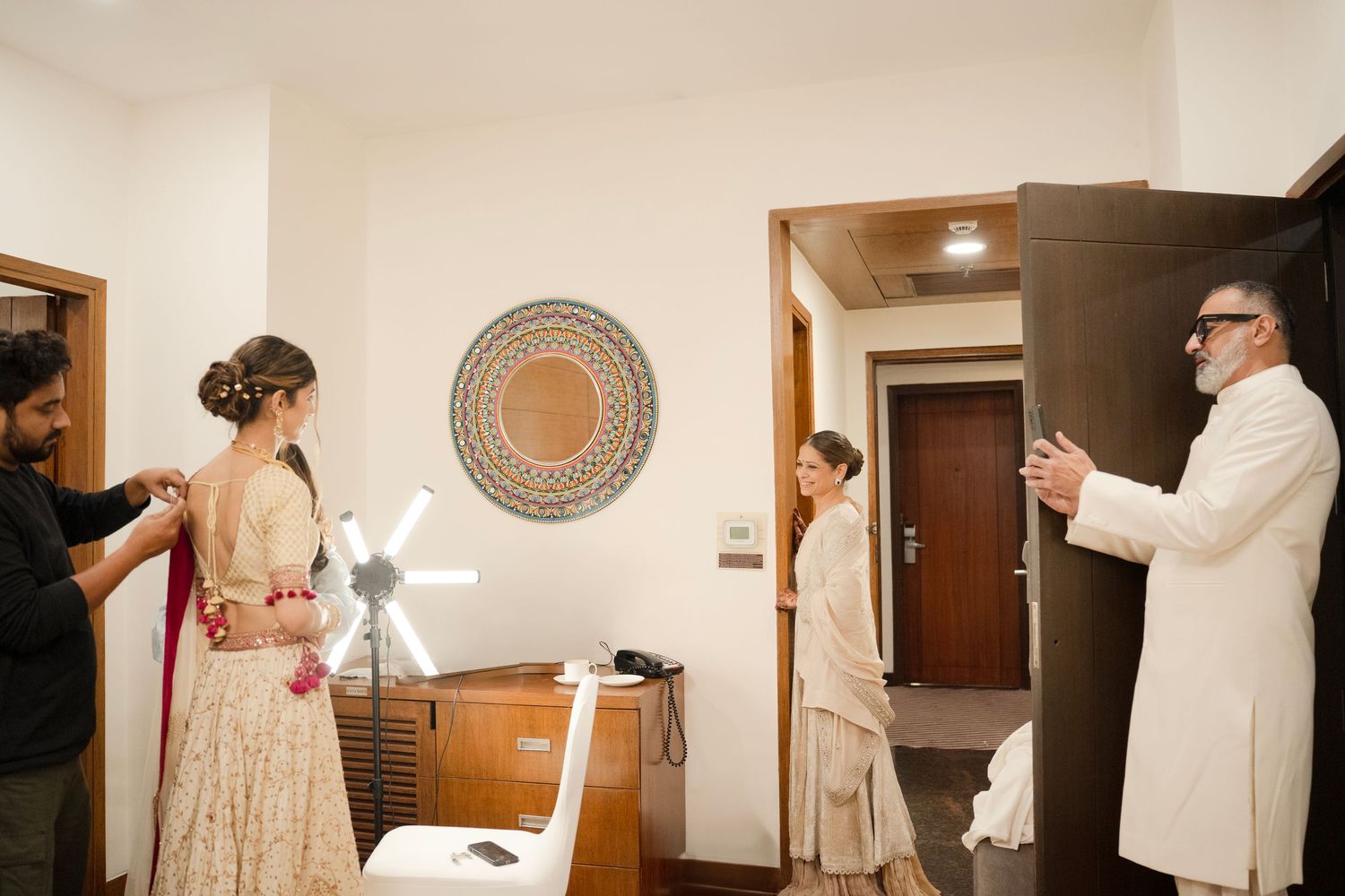 Bride getting ready in hotel room with family members watching during Indian wedding preparations.