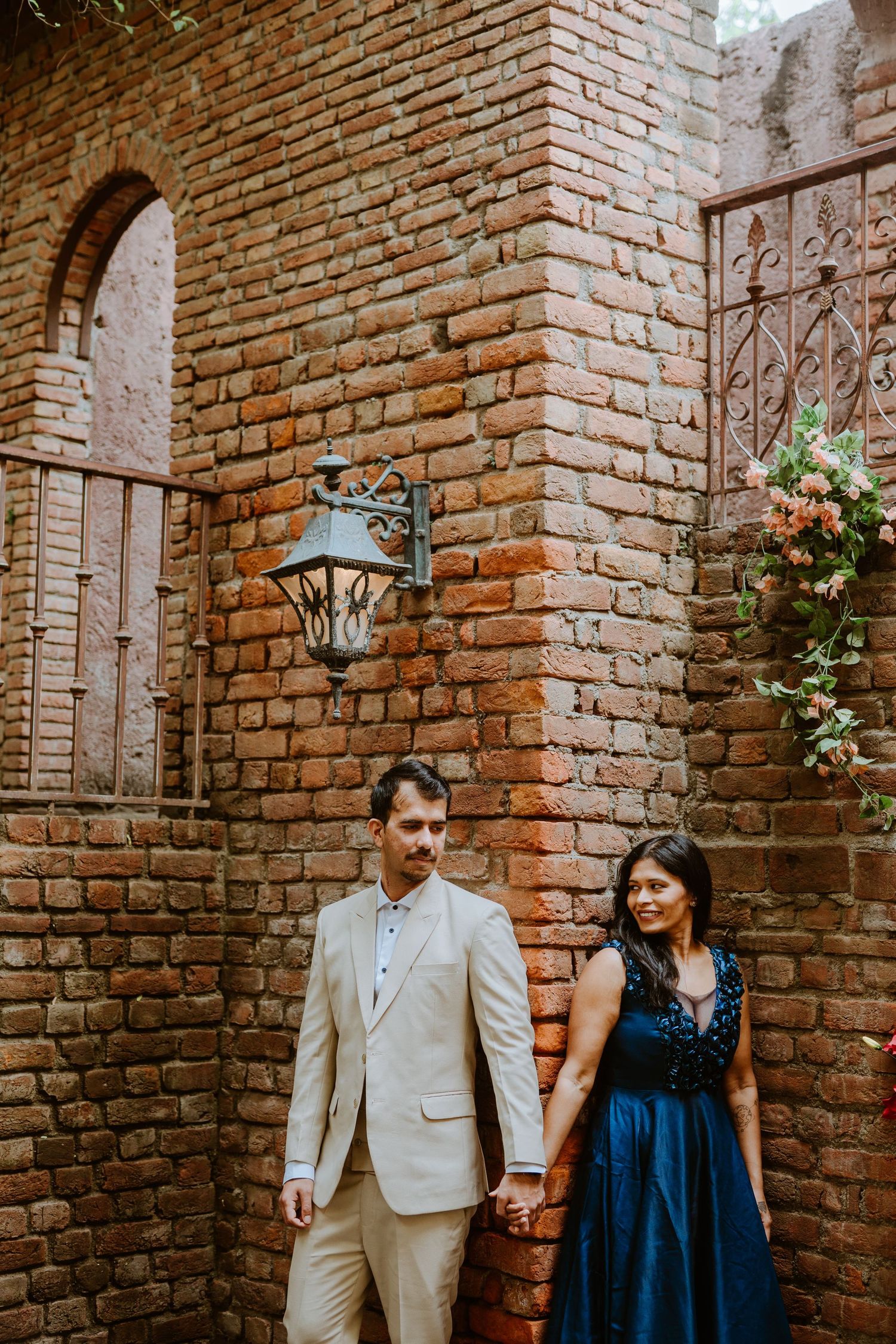 Couple holding hands in front of rustic brick wall with vintage lantern, man in beige suit, woman in navy dress.