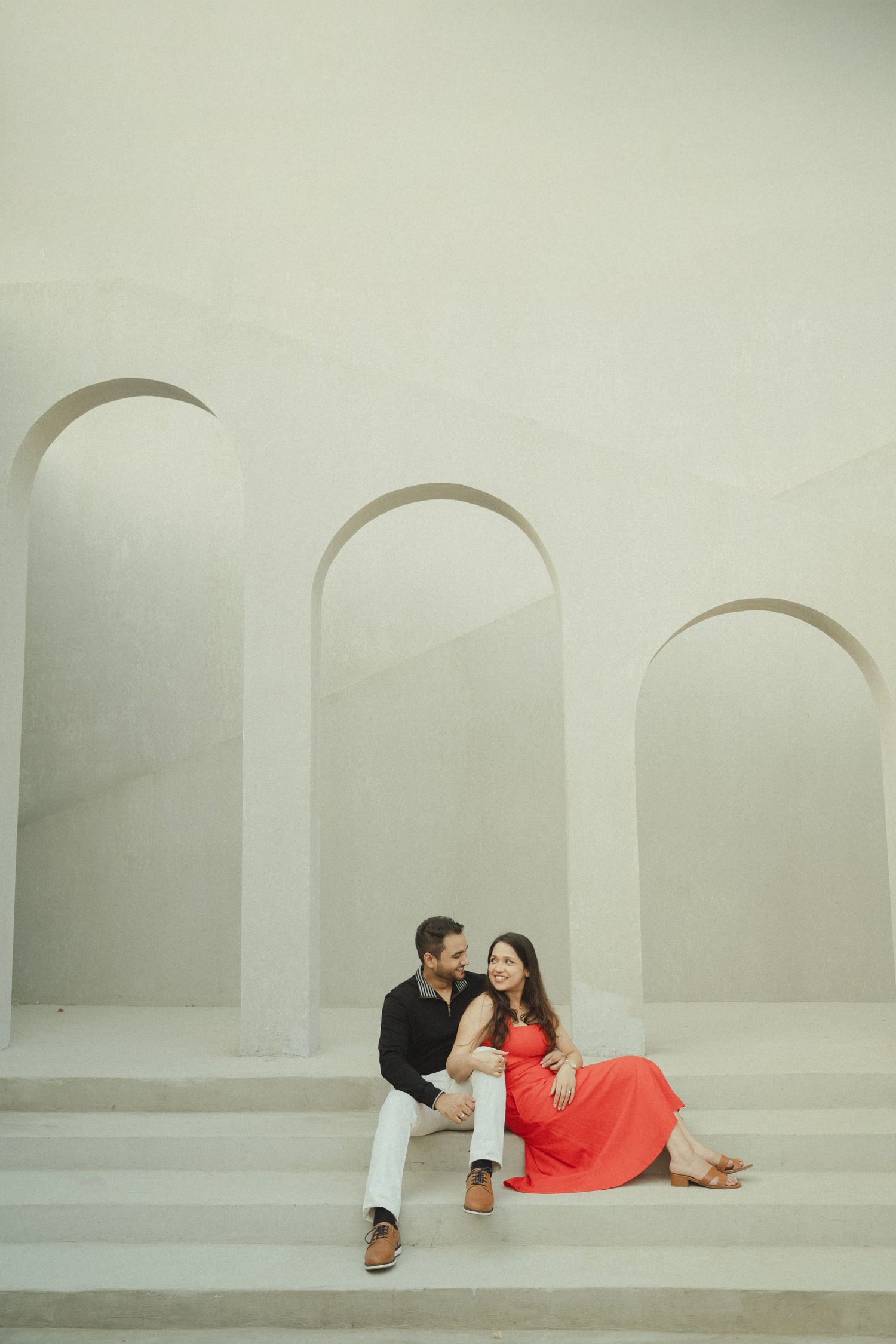 Couple sitting on white steps with arched walls, woman in red dress, man in black shirt and white pants.
