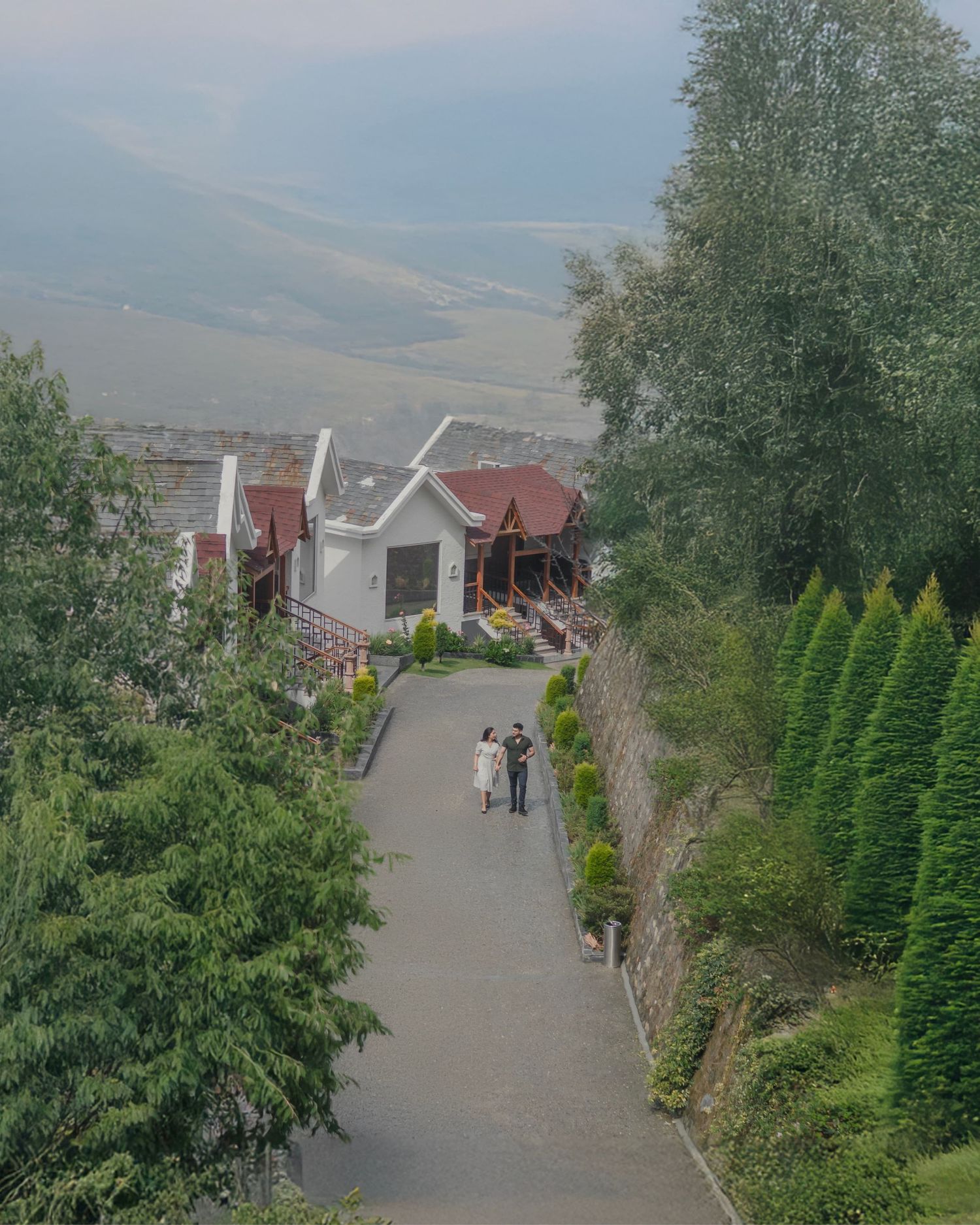 Two people stroll down a paved path flanked by lush trees and red-roofed buildings overlooking a misty valley.