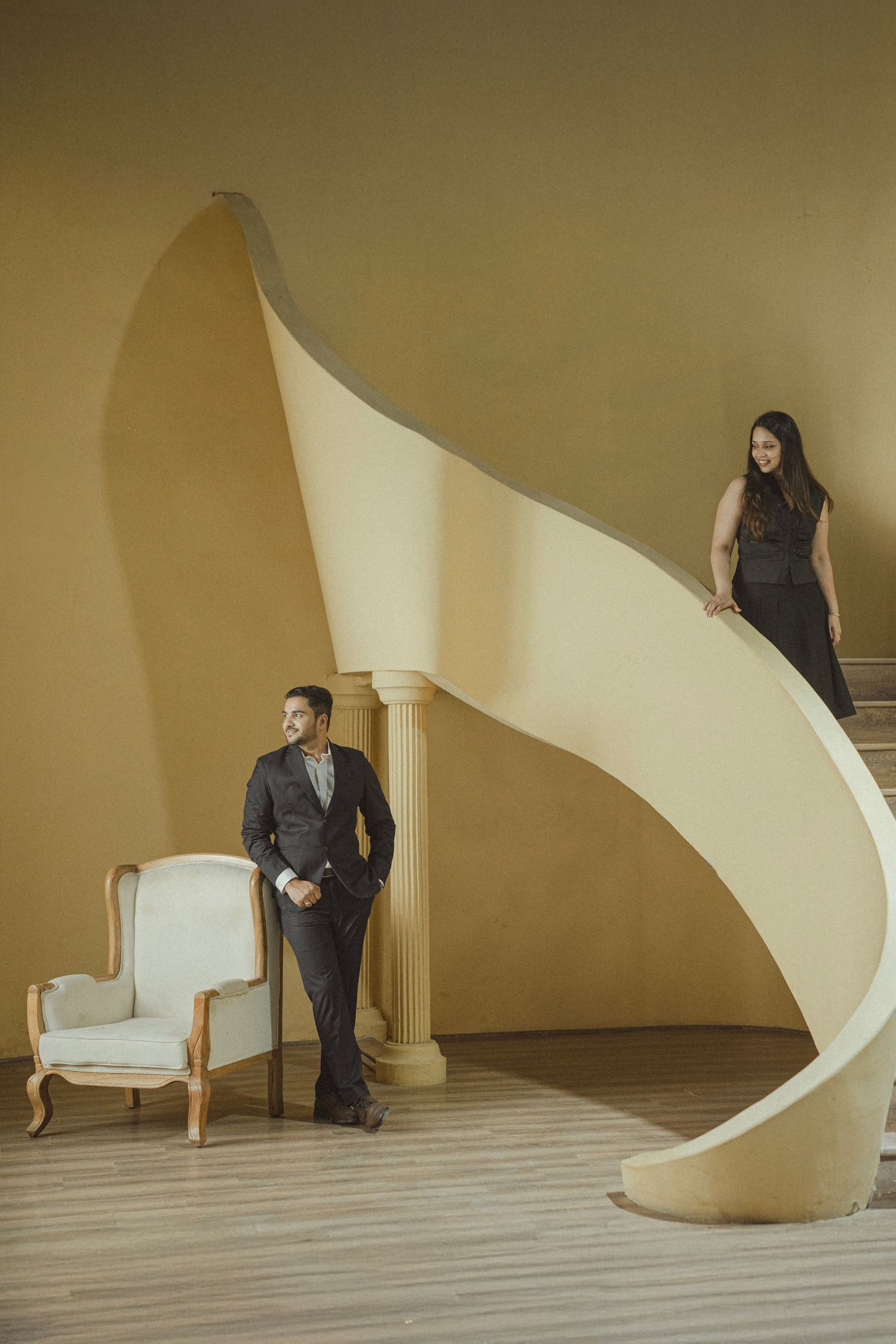 Elegant couple in formal attire posed on a curved staircase in a golden-hued architectural interior.