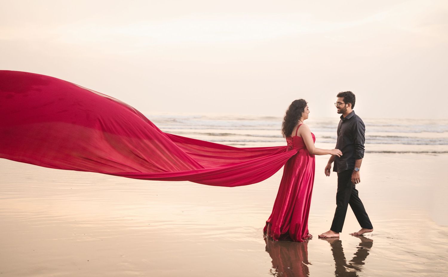 A flowing red dress creates dramatic movement during a beach photoshoot at sunset.