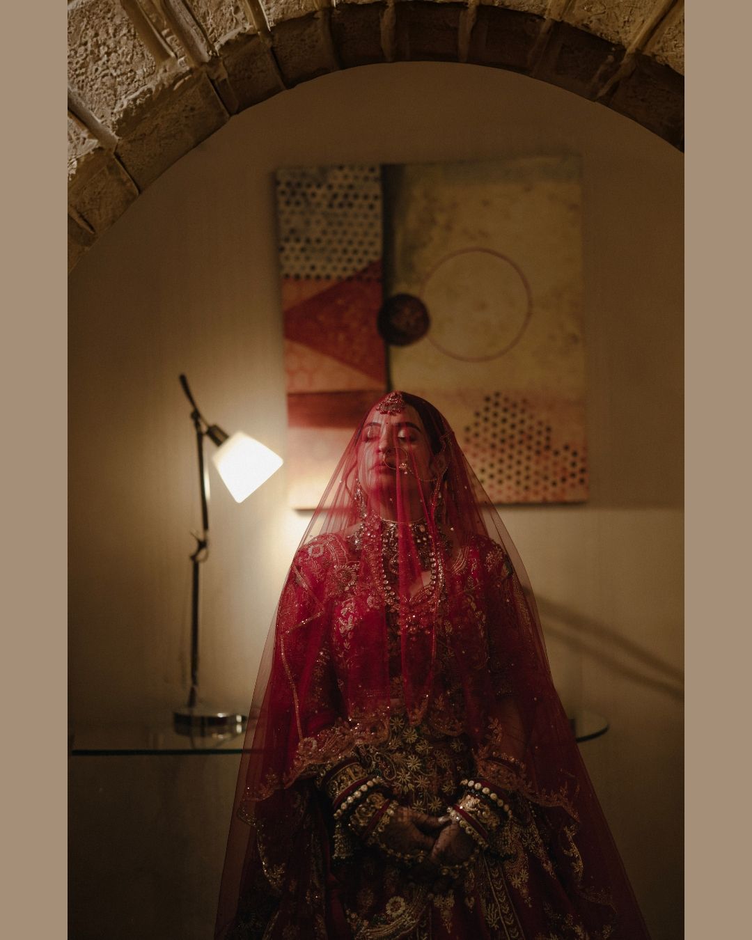 A bride in a red lehenga and dupatta veil stands under an arch in moody lighting.