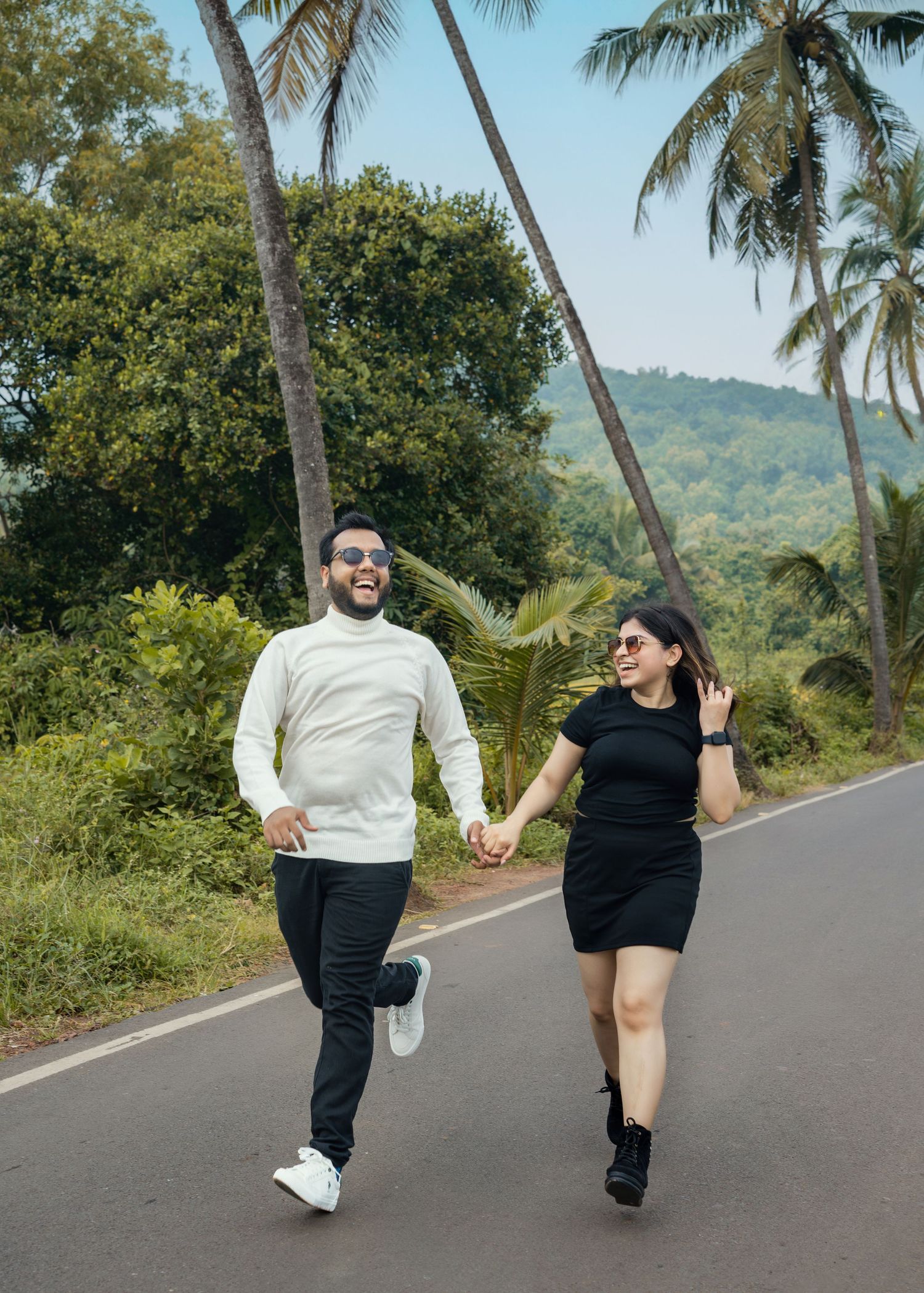 Couple running playfully on a palm tree lined road with mountains in the background.
