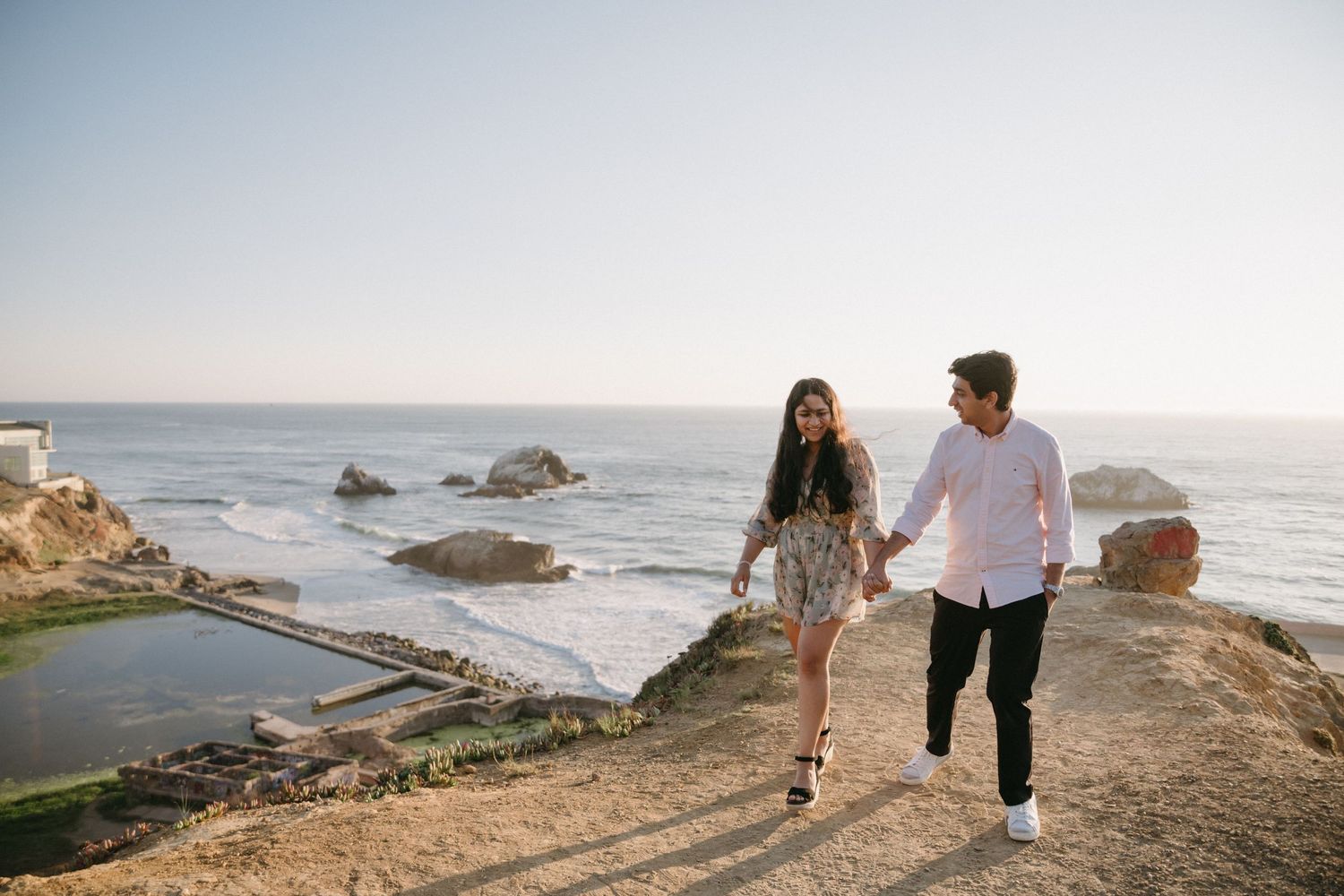 A couple walks along coastal cliffs at sunset during their engagement shoot.