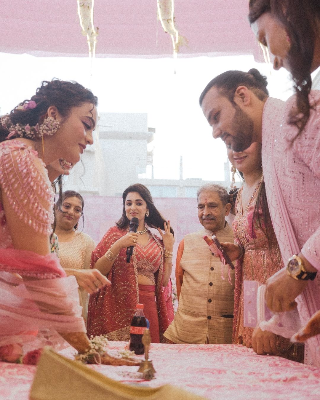 A joyful Indian wedding ceremony with guests in pink traditional attire gathered around a ritual table indoors.