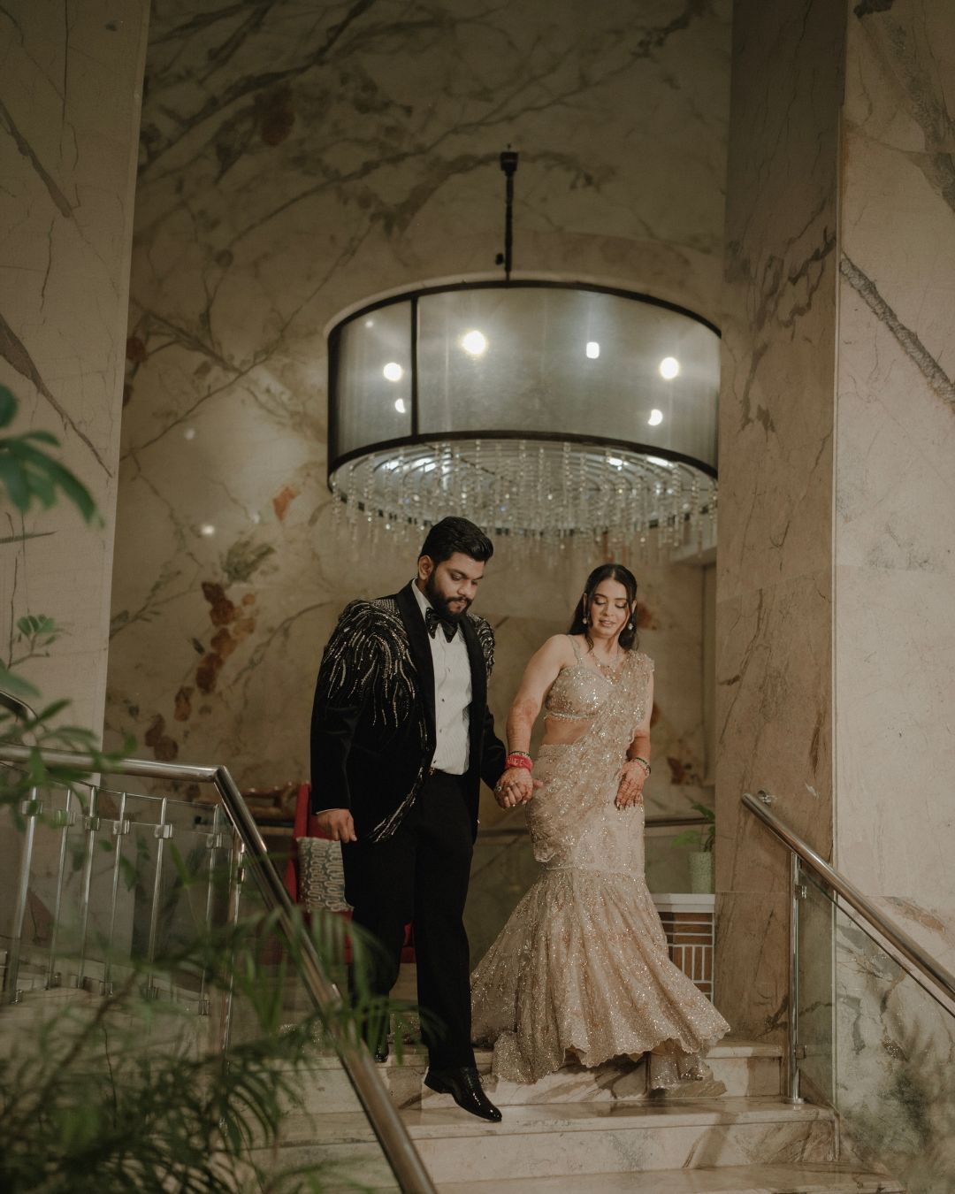 Elegant couple descending marble staircase under crystal chandelier, groom in black tuxedo, bride in gold embroidered saree gown.