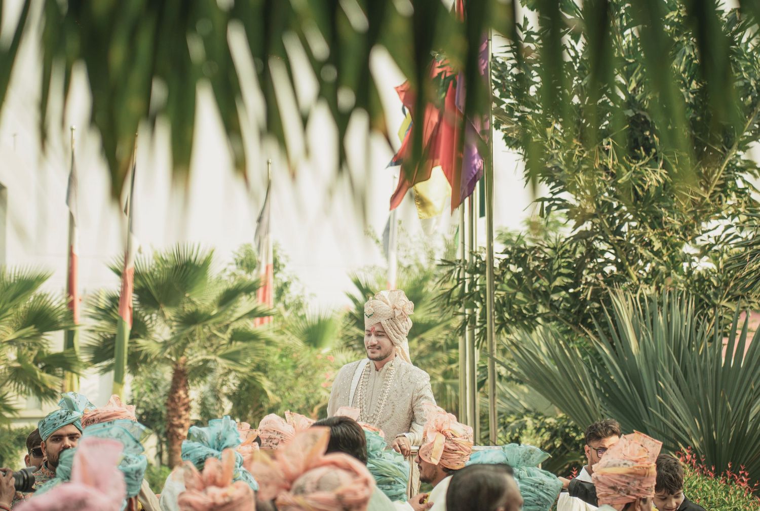 Groom in traditional attire stands among colorfully dressed wedding guests surrounded by palm trees and festive flags.