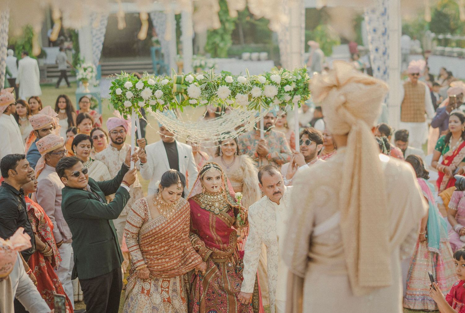 Indian bride walks down the aisle under floral canopy at traditional Hindu wedding ceremony with guests.