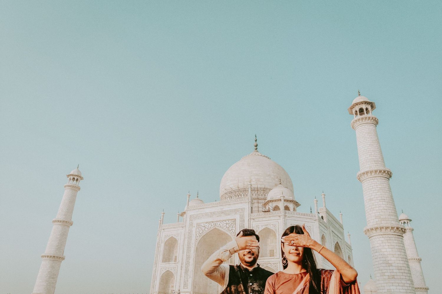 Couple posing playfully in front of the iconic Taj Mahal in Agra, India under a clear blue sky.
