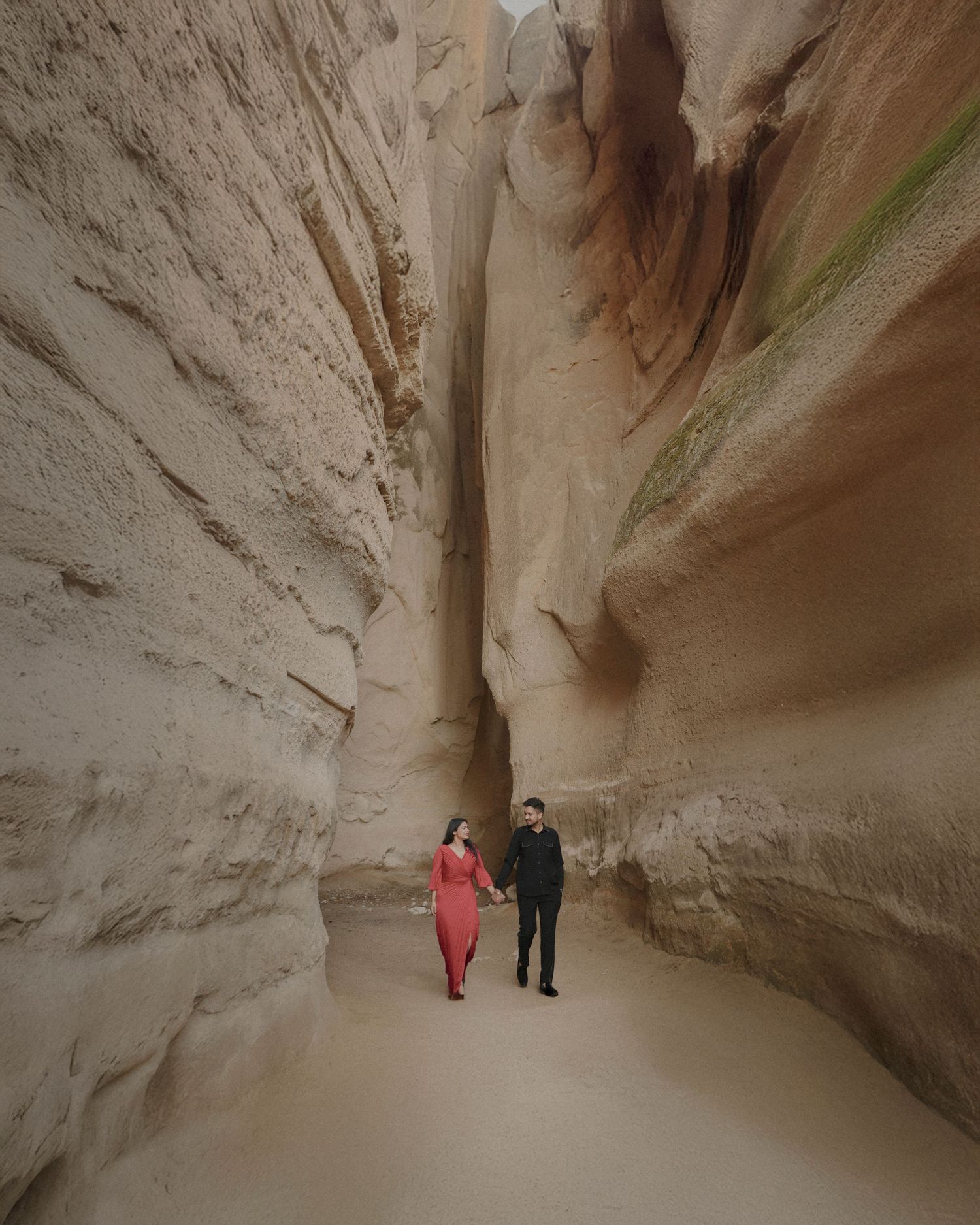 Couple holding hands walking through a narrow slot canyon with towering sandstone walls, woman in red dress.