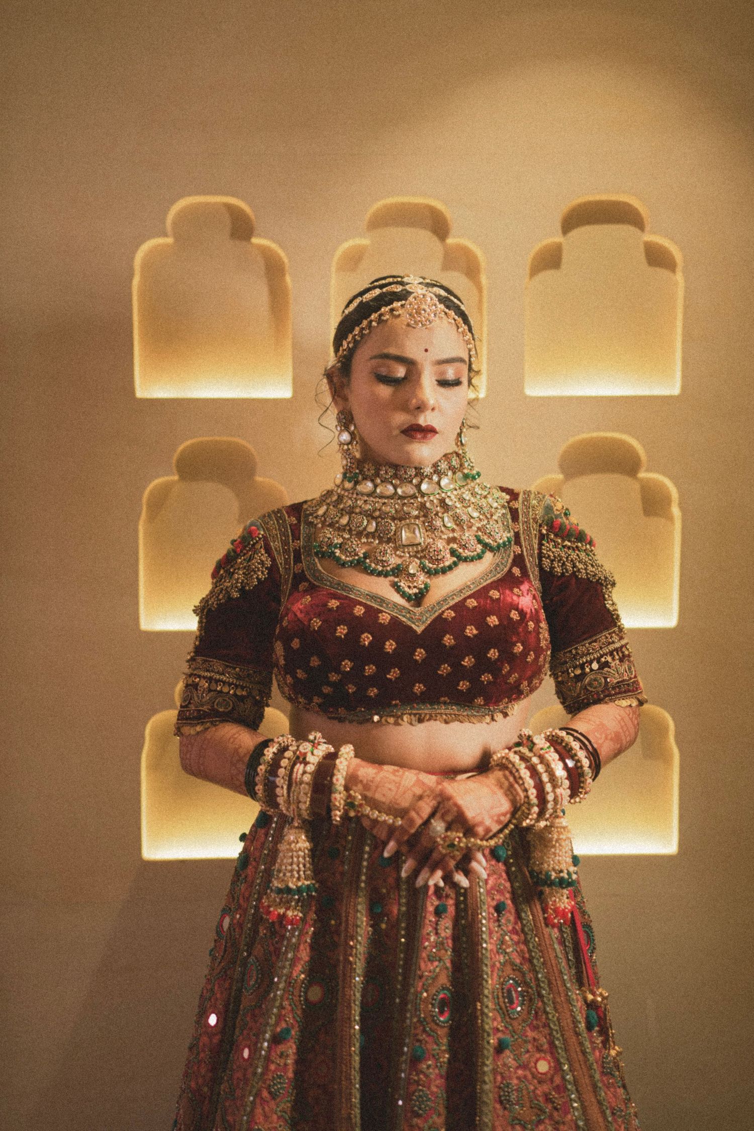 Indian bride in ornate maroon lehenga with gold jewelry and maang tikka poses gracefully against illuminated wall backdrop.