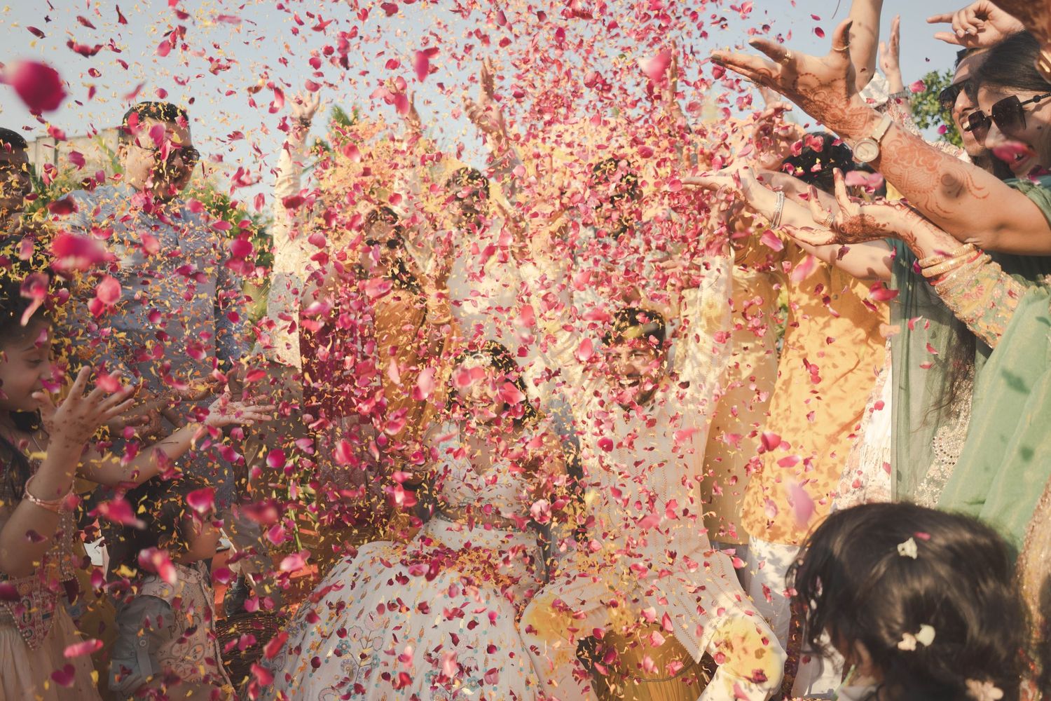 Joyful wedding celebration with guests throwing colorful flower petals and confetti over the bride and groom outdoors.