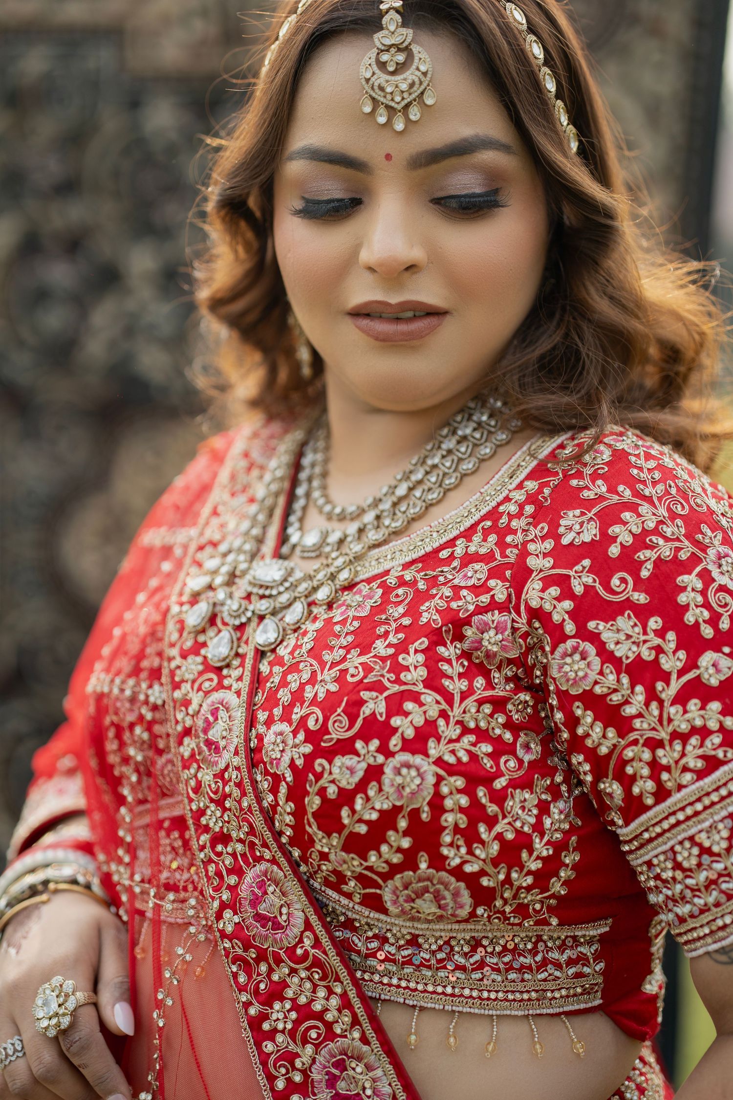 A bride in a red embroidered lehenga with gold jewelry, maang tikka, and traditional bridal makeup looks down gracefully.