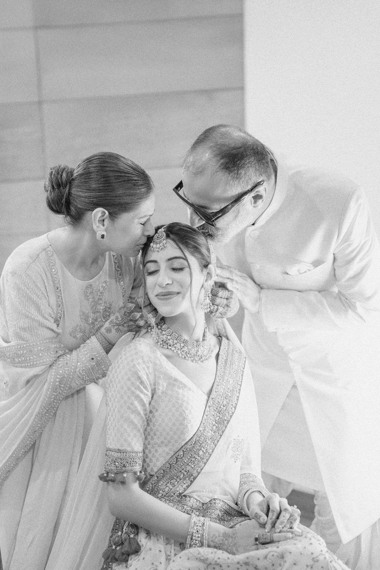 Black and white photo of a bride in traditional Indian attire being blessed by her parents on her wedding day.