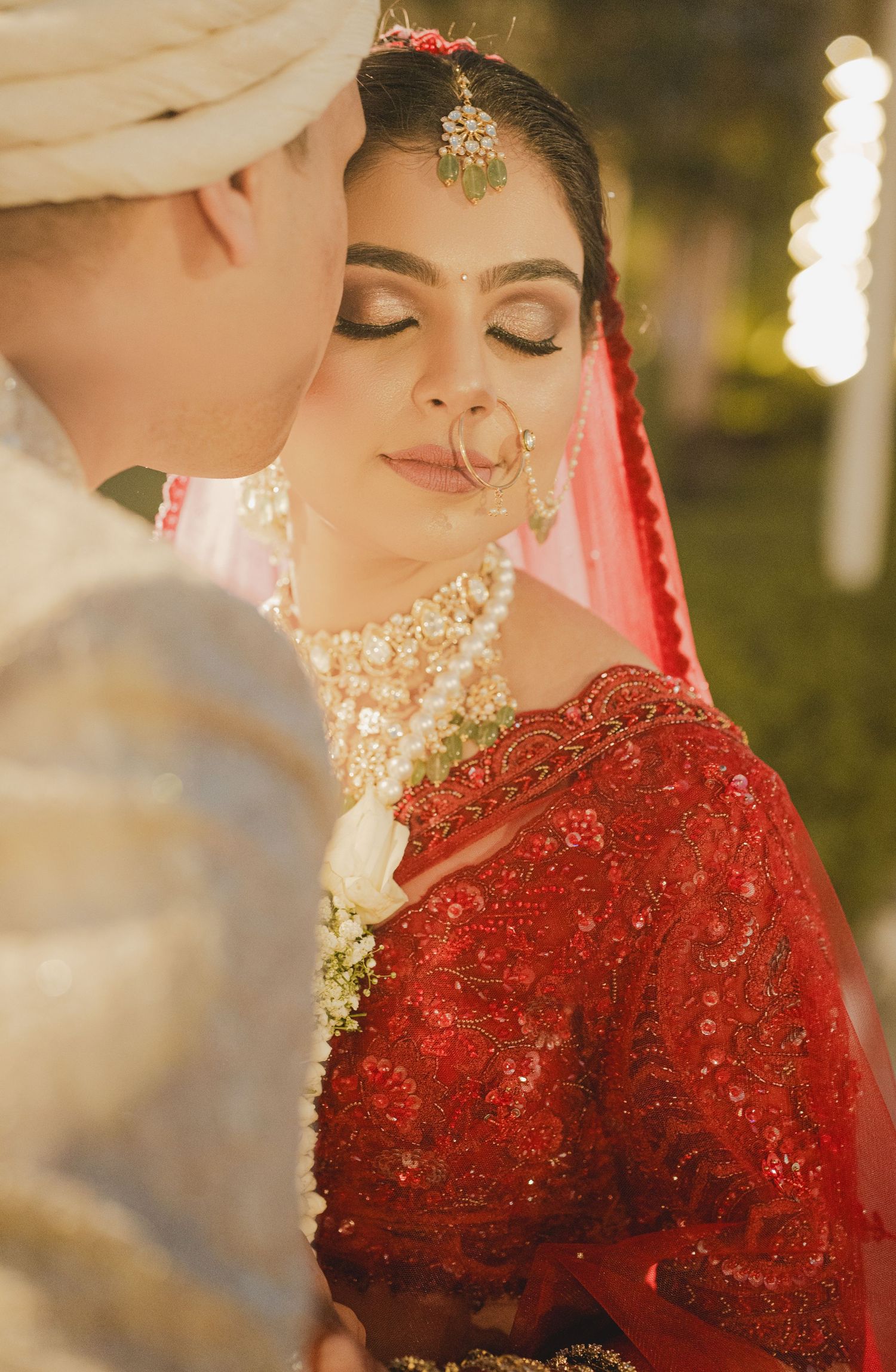 Traditional South Asian wedding attire featuring an ornate red lehenga with pearl necklace and golden tikka jewelry.