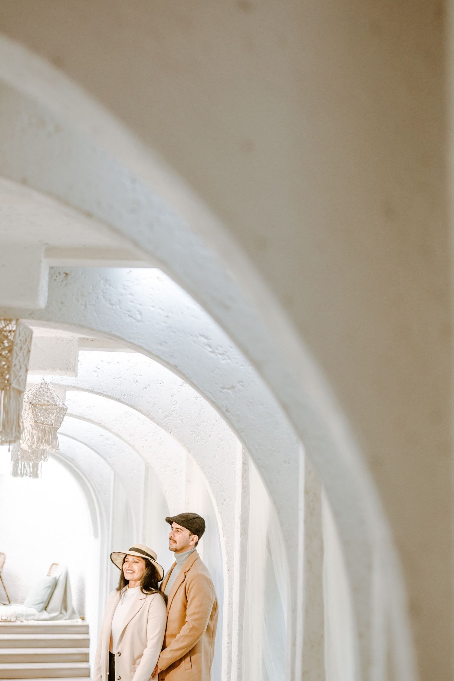 Couple in beige outfits standing under white arched corridor, woman in sun hat, man in cap, romantic architectural setting.