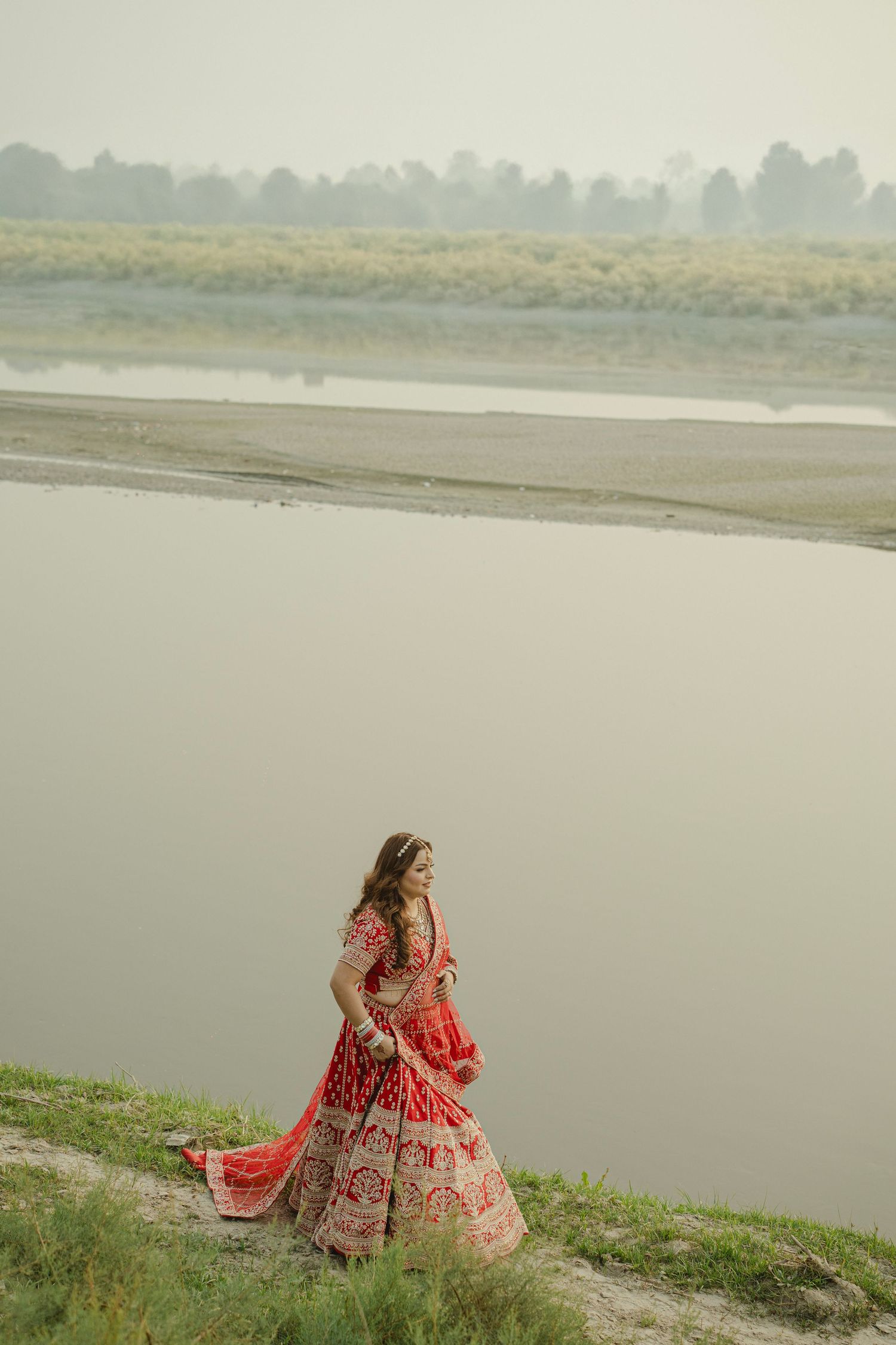 Bride in red lehenga walking by a serene river, wearing traditional Indian bridal attire with intricate embroidery.