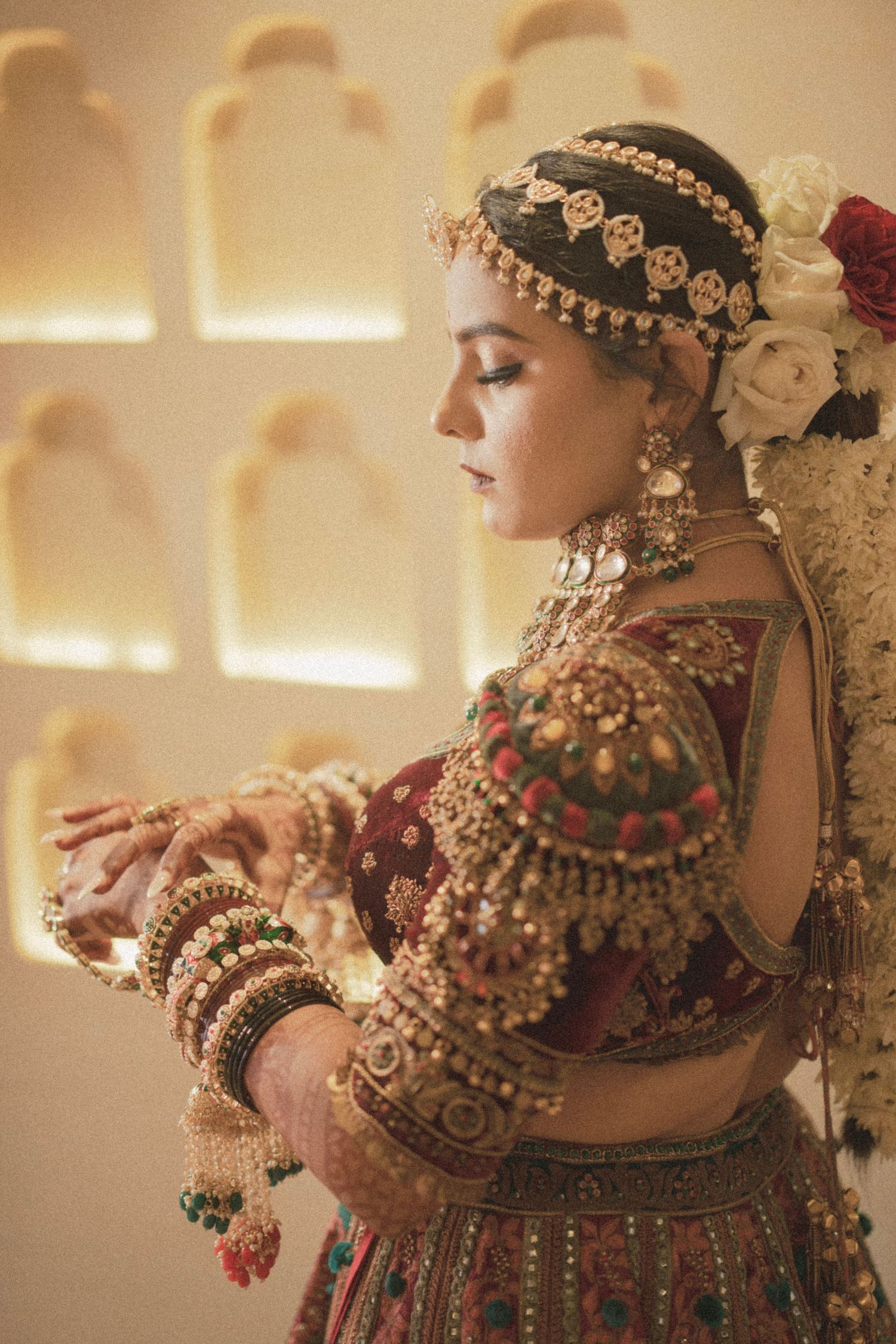 Indian bride in red lehenga with gold jewelry, maang tikka, and floral hair accessory adjusting bangles.