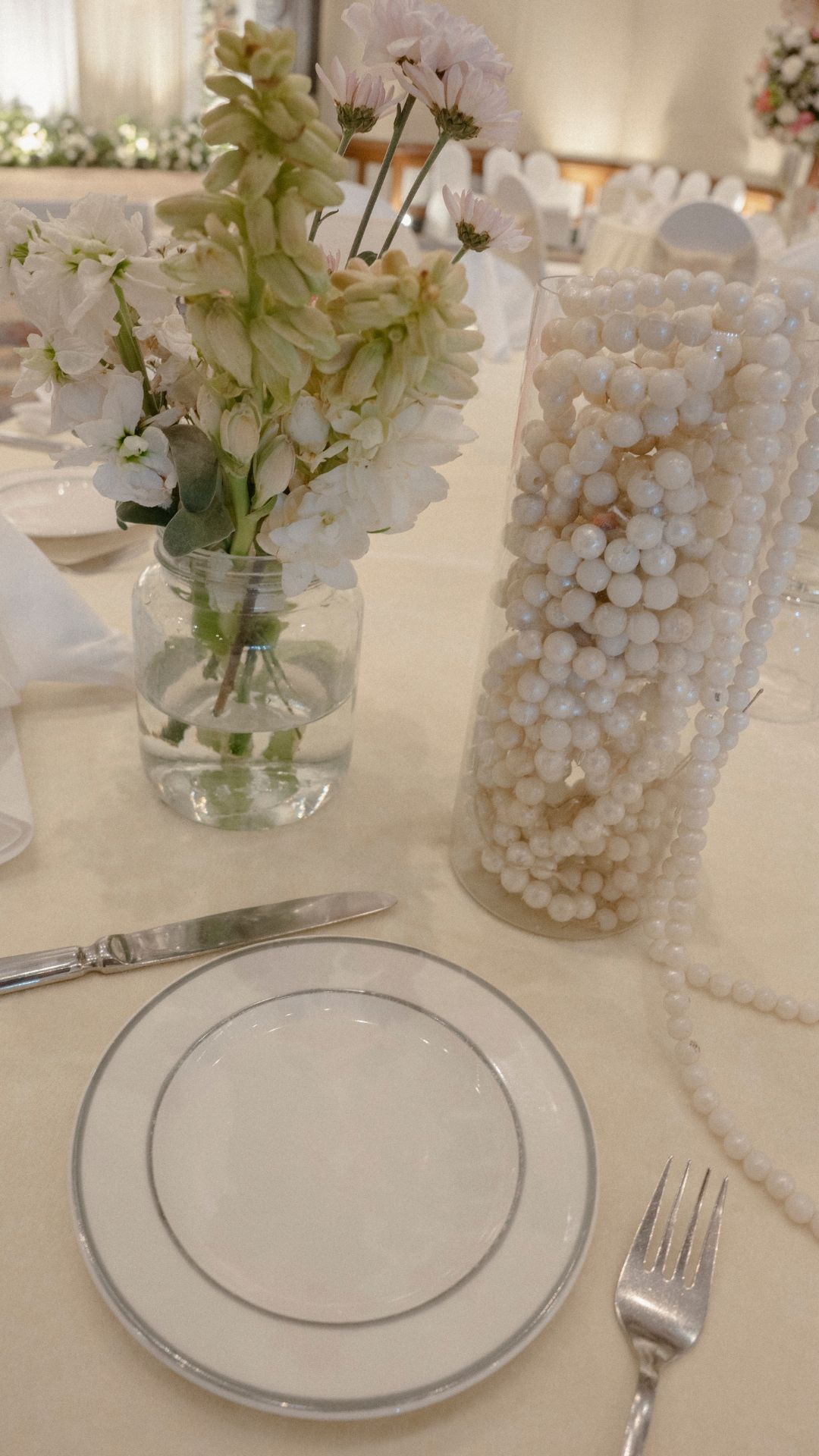 Elegant white wedding table setting with pearl vase, mason jar florals, silver-rimmed plate, and silverware on ivory linen.