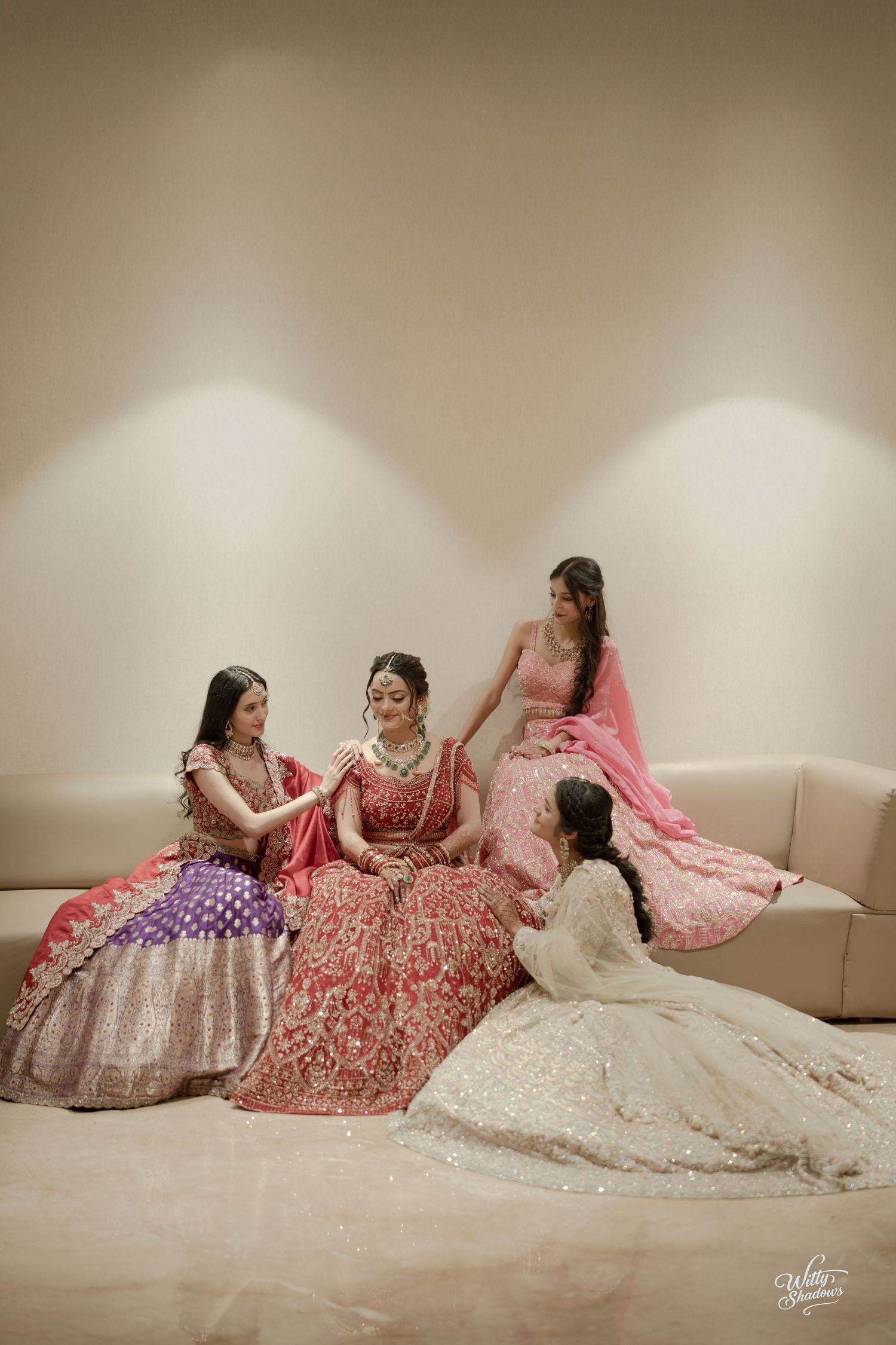 Four women in elegant Indian wedding attire including lehengas in red, pink and cream colors sit together on a beige couch.
