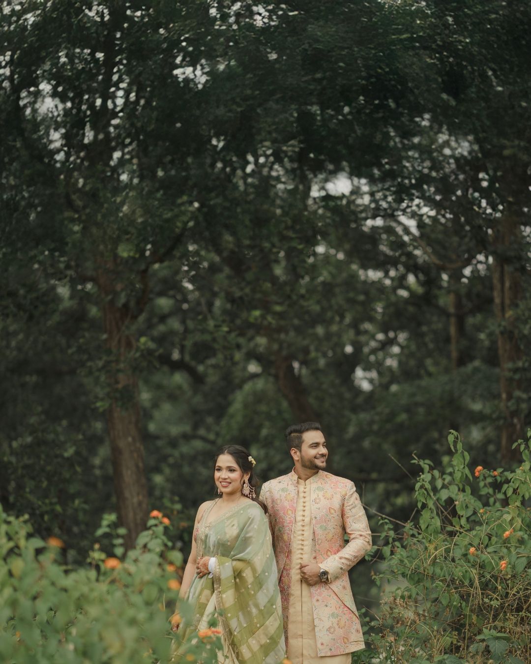 Couple in traditional Indian attire walking through lush green garden, woman in saree, man in sherwani.