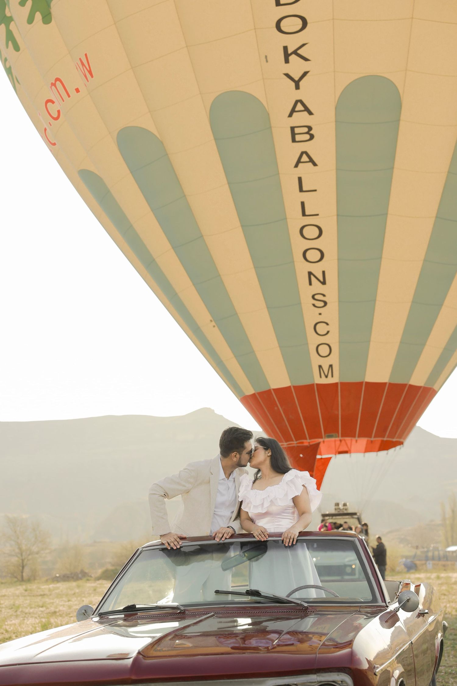 Couple sharing a romantic kiss standing in a convertible car with a colorful hot air balloon behind them.