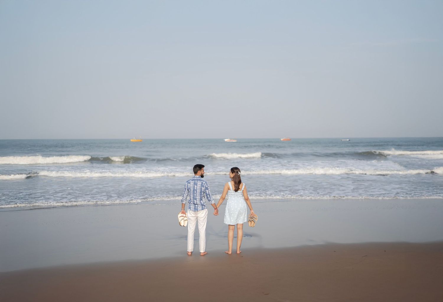 Couple holding hands on a sandy beach, facing ocean waves with ships visible on the horizon.