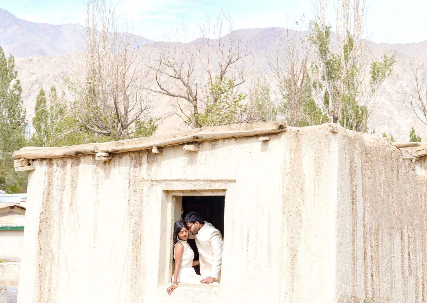 Couple sharing a romantic moment in a rustic adobe window with mountain backdrop and bare trees.