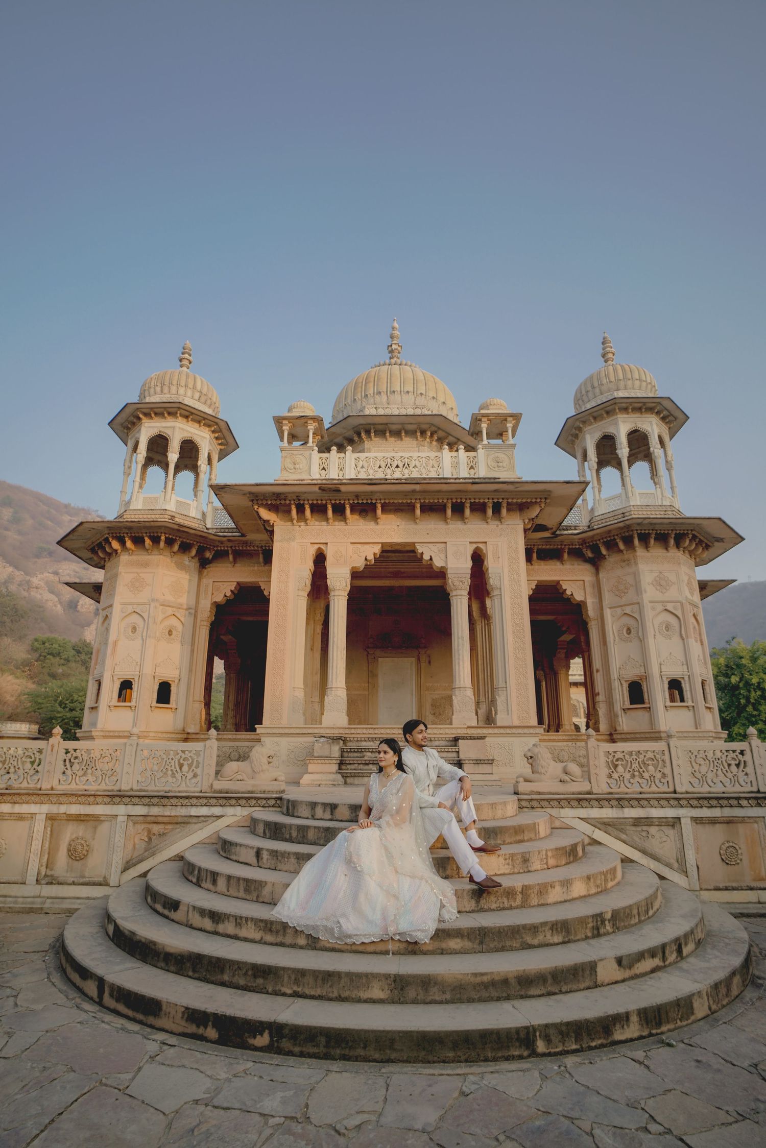 Couple in wedding attire posing on steps of ornate Rajasthani palace with domed architecture at dusk.