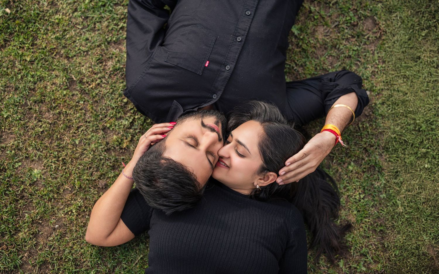 Couple in black outfits lying on grass sharing a romantic kiss in an outdoor setting.