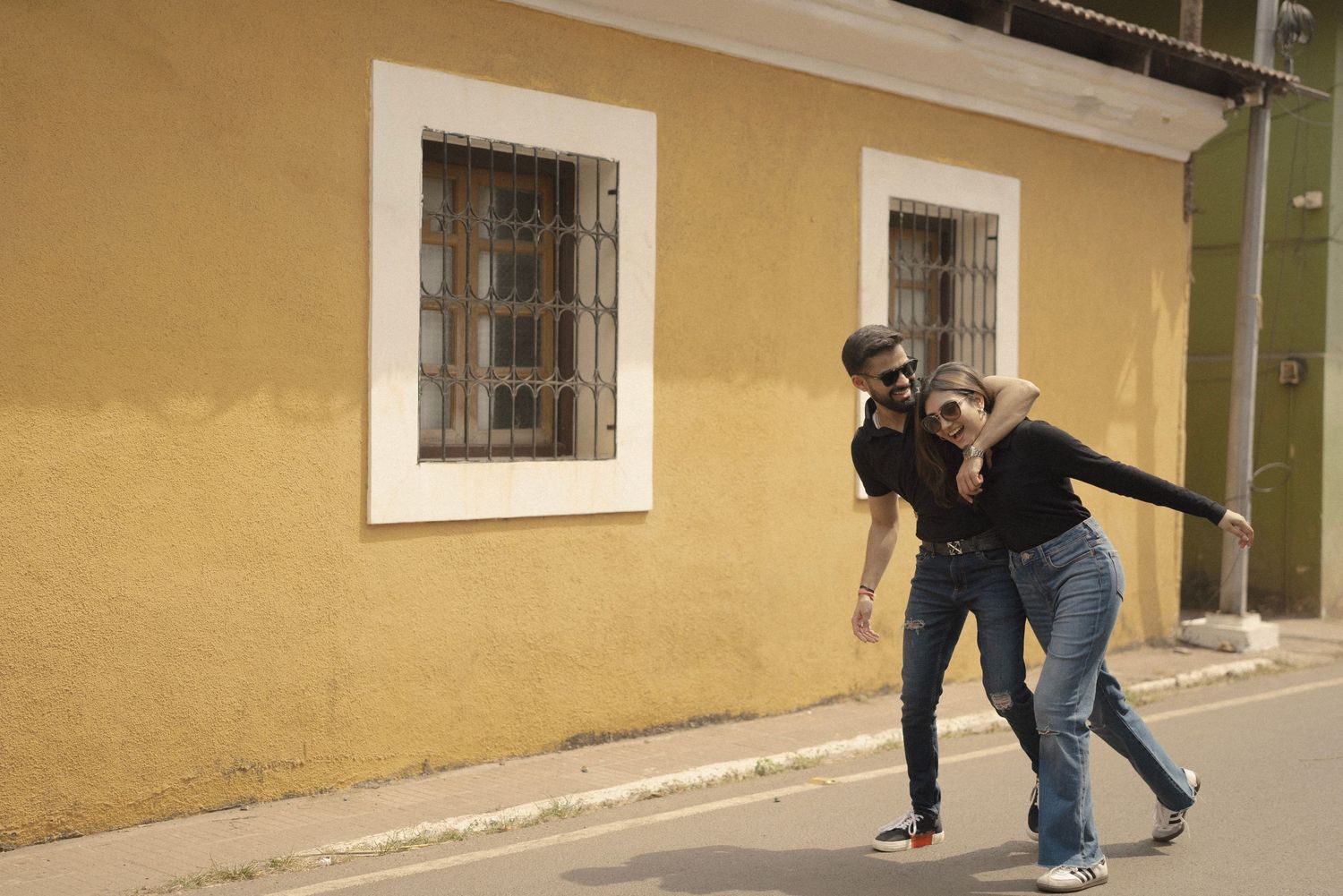 A cheerful couple laughing and posing playfully on a street beside a yellow building with barred windows.